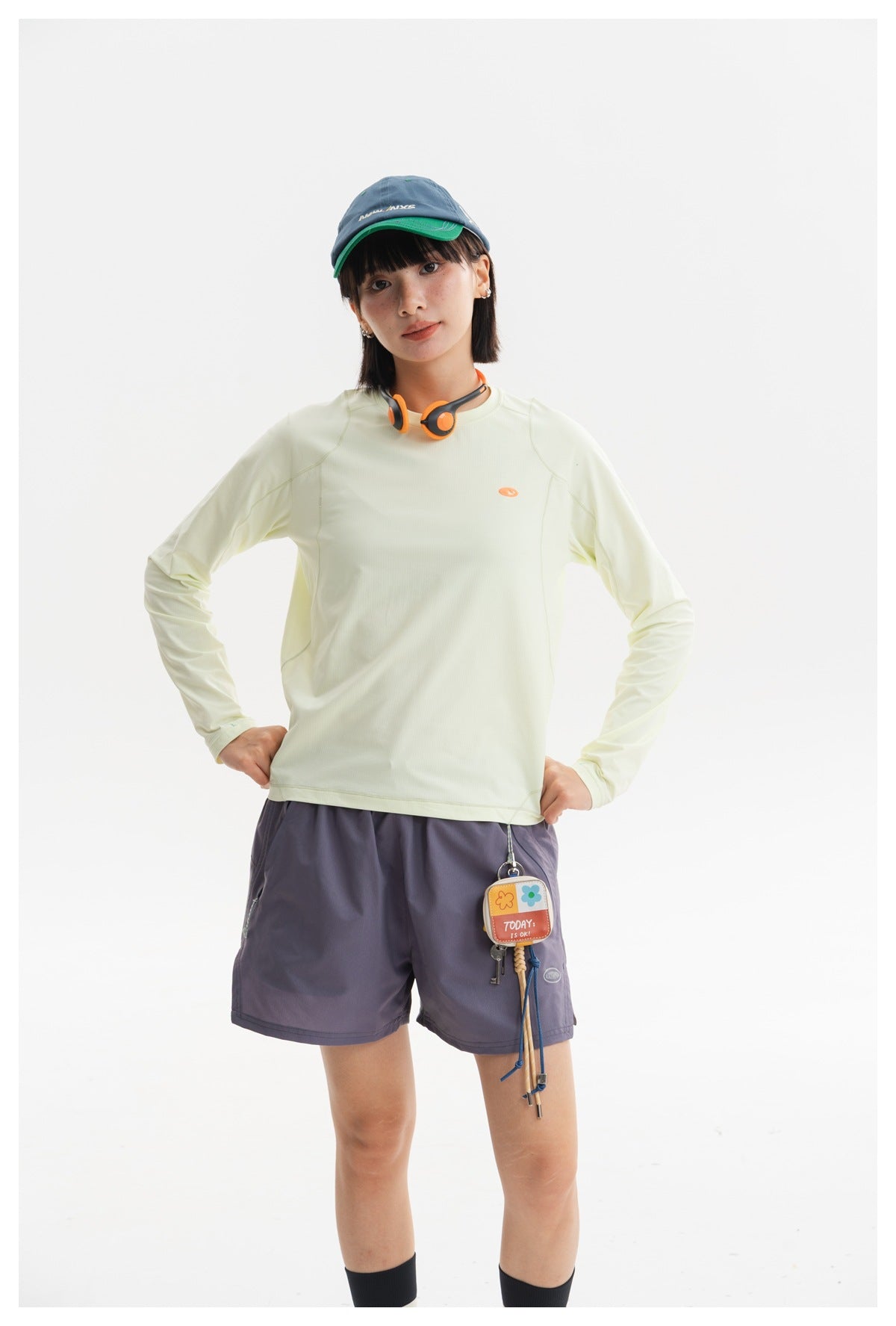 Model wearing a pale yellow-green long-sleeve top and slate-purple shorts, standing front view.