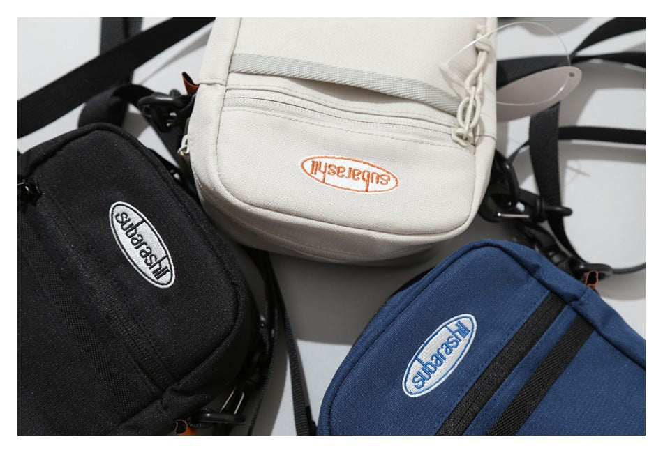 Black, cream, and navy crossbody bags arranged together, showing zip pockets, straps, and oval embroidered labels.