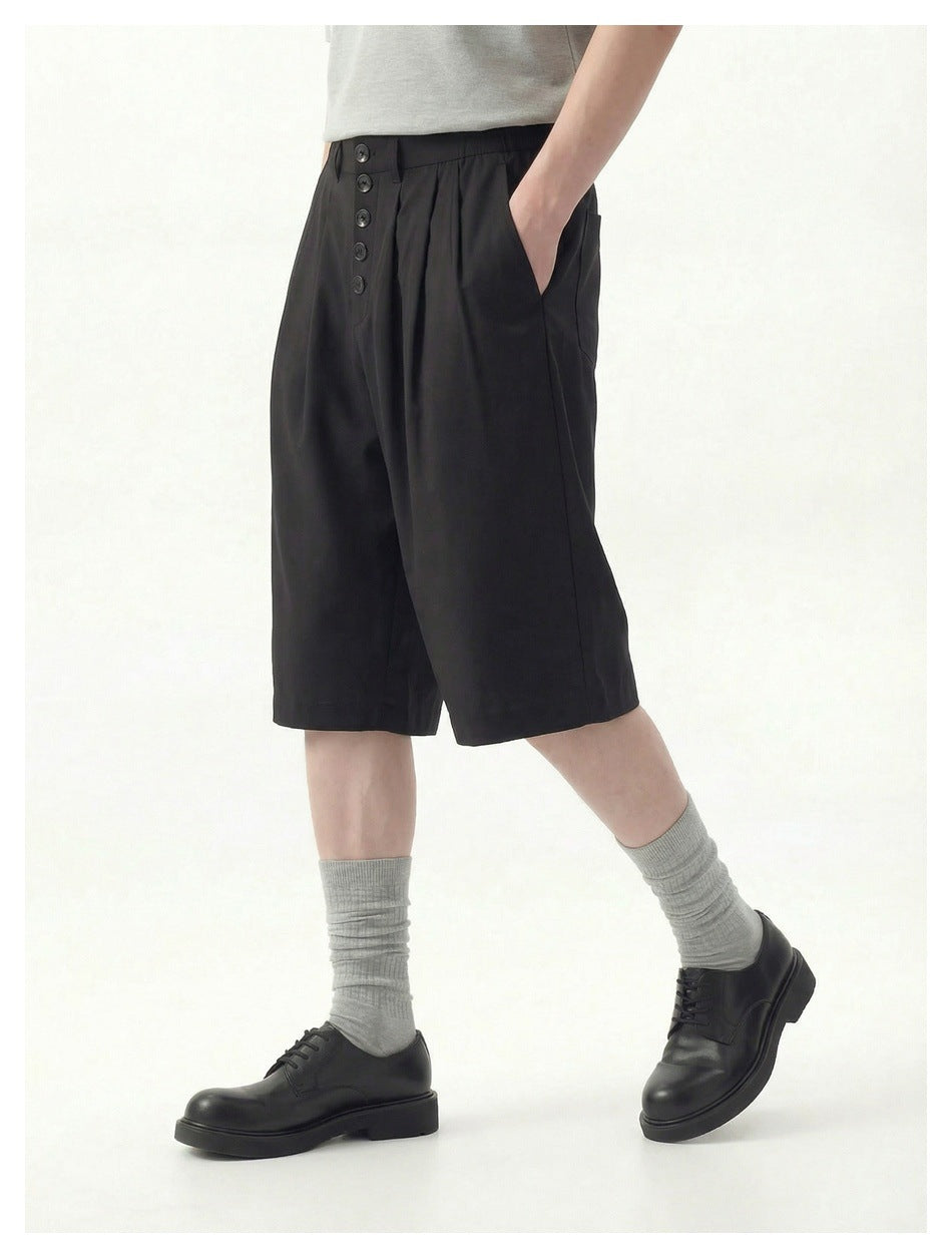 Model wearing black pleated knee-length shorts with button front, gray socks and black leather shoes, hand in pocket