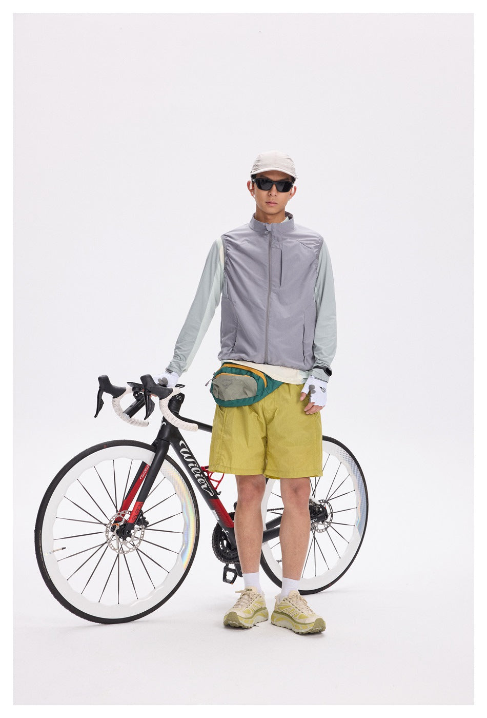 Cyclist wearing a gray zip-front vest over a pale mint long-sleeve shirt and mustard-yellow shorts, standing beside a bike.