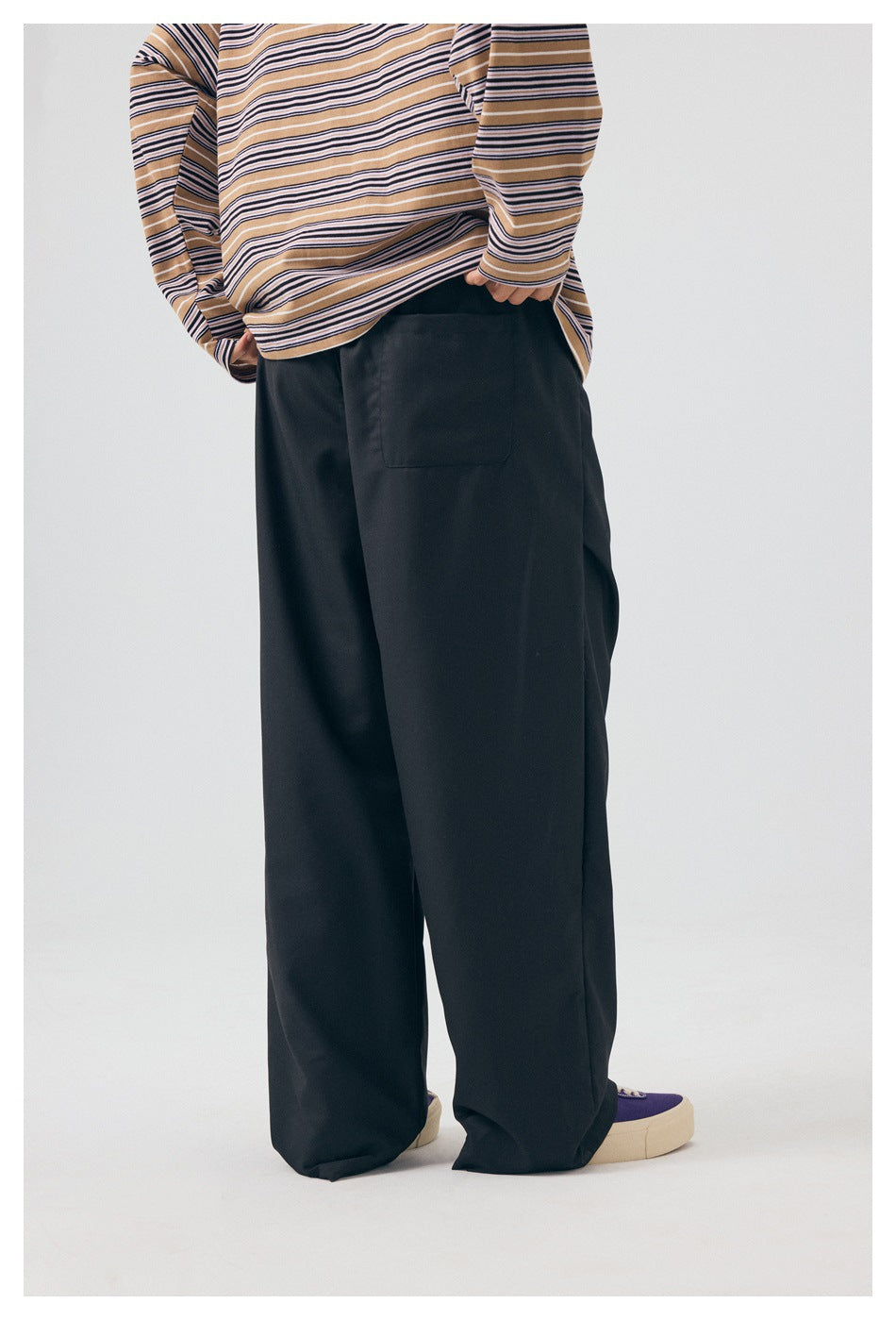 Rear view of a model wearing loose black trousers with a back pocket and a tan striped long-sleeve top.