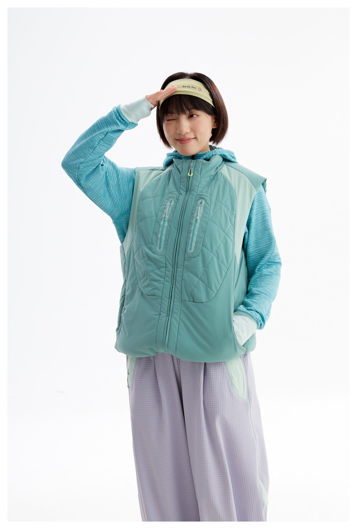 Closer front view of a model wearing a teal quilted zip vest with a hood and two zippered chest pockets.