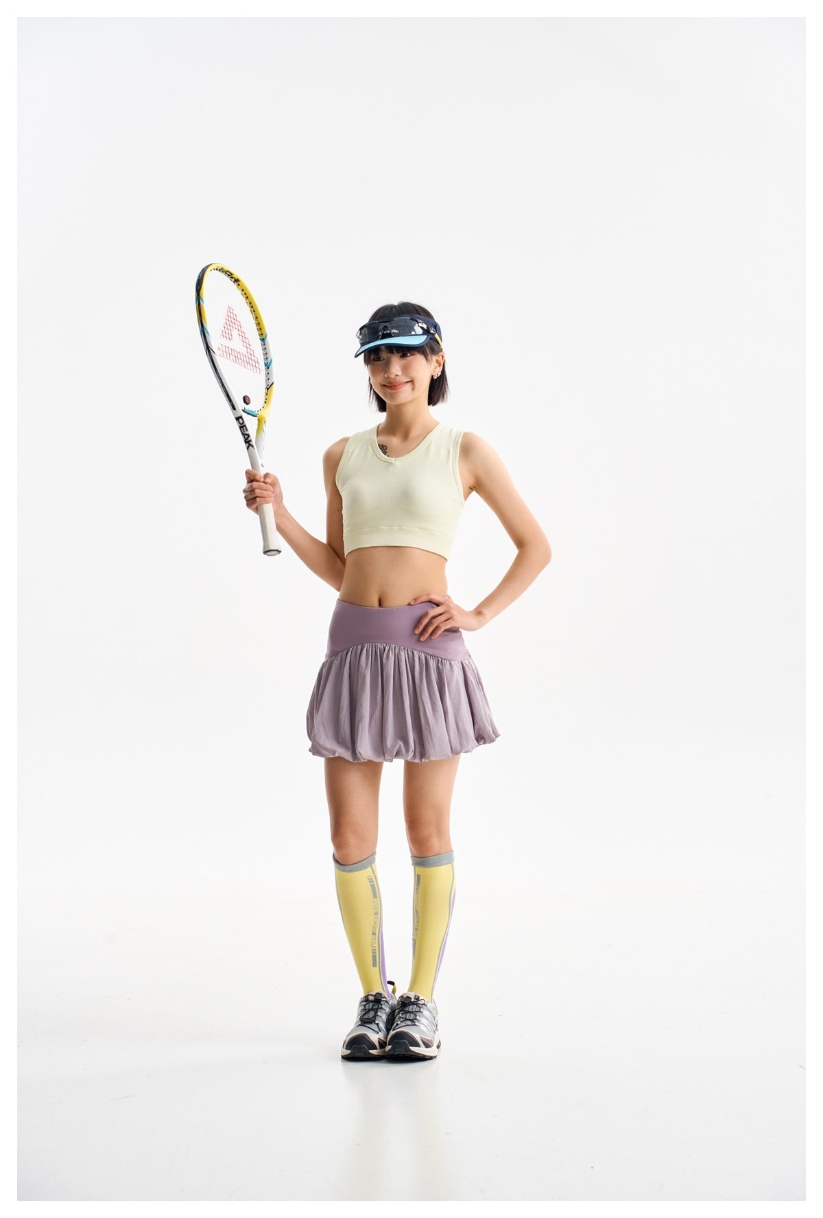 Model holding a tennis racket, wearing a pale yellow cropped top and lavender-gray bubble-hem skirt.