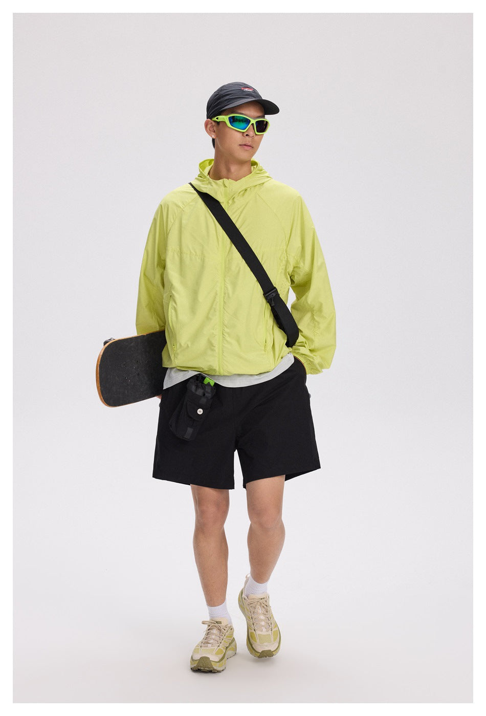 A model wears a lime hooded jacket with black shorts and carries a skateboard and a black crossbody bag.