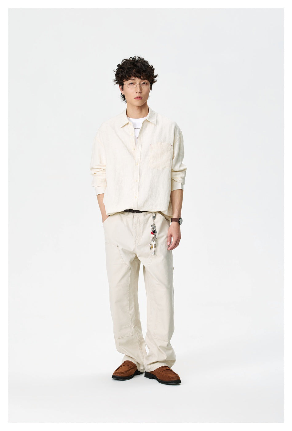 Man standing in an off-white button-up shirt over a white tee with cream trousers and brown loafers