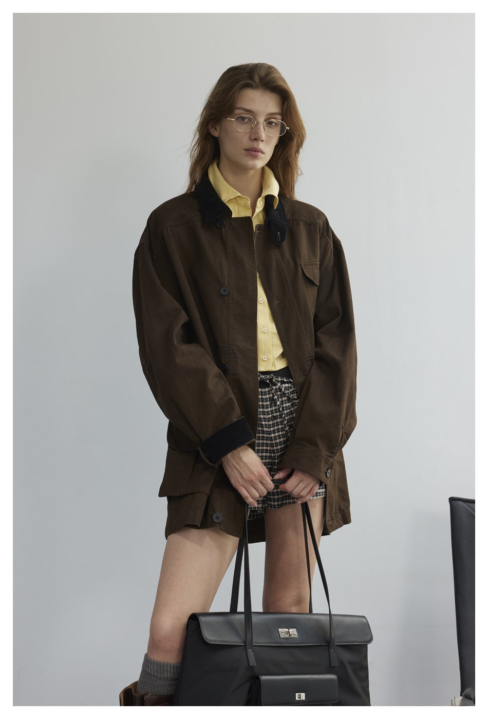 Woman in a brown jacket with black corduroy-look collar and cuffs, yellow shirt and plaid shorts, holding a black tote