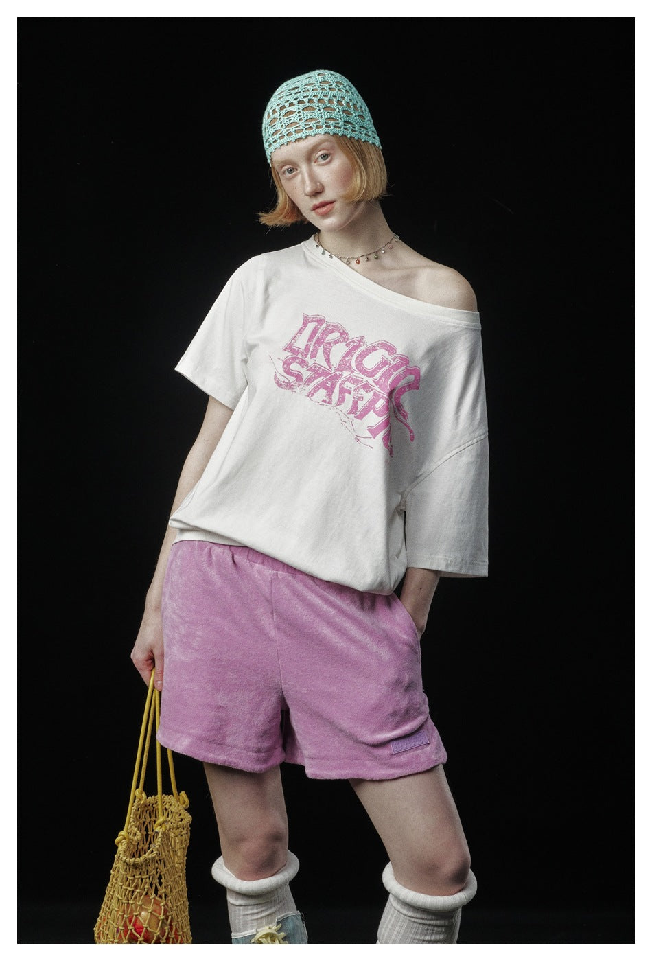 Closer view of a model in an off-white, pink-graphic T-shirt and pink shorts, with a turquoise open-knit cap.