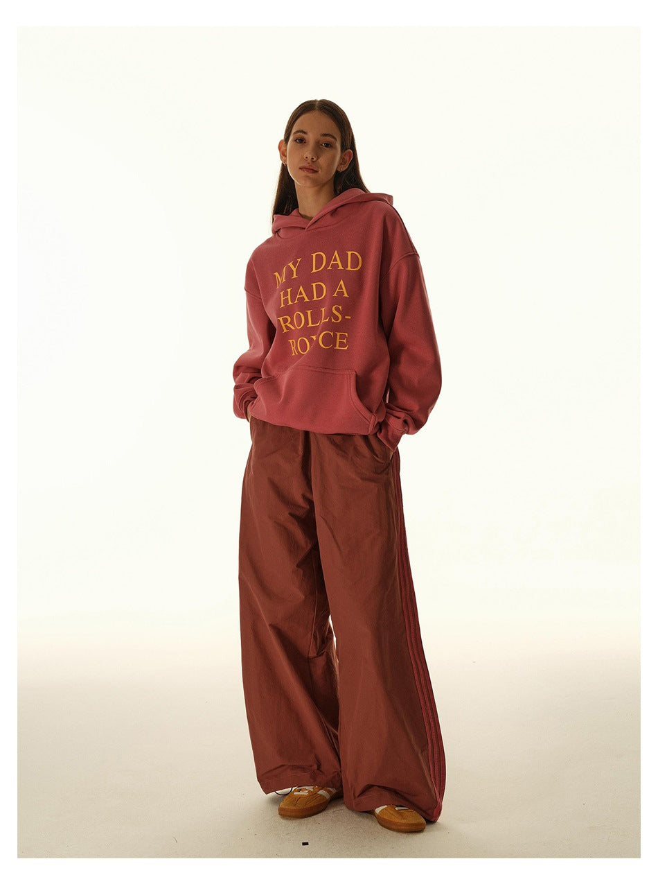Model wearing a rose-pink hoodie with gold lettering and wide rust-brown pants with side stripes.