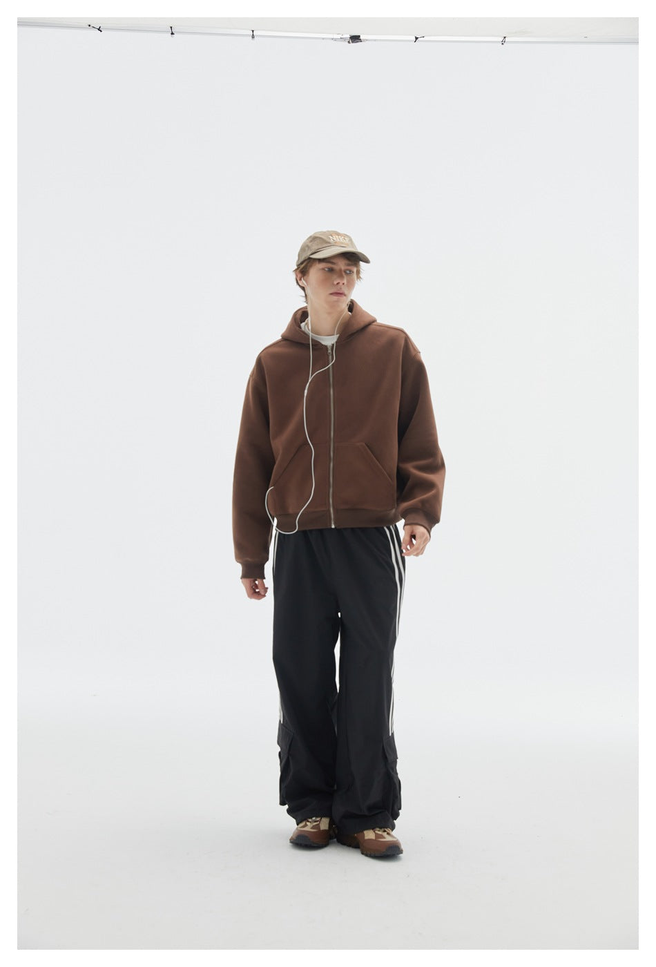 Model wearing a brown zip-up hoodie with black striped cargo pants and a cap