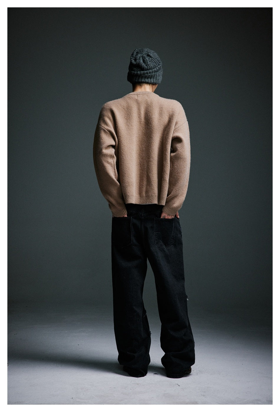 Back view of a model wearing a plain tan sweater, loose black jeans, and a dark ribbed beanie.