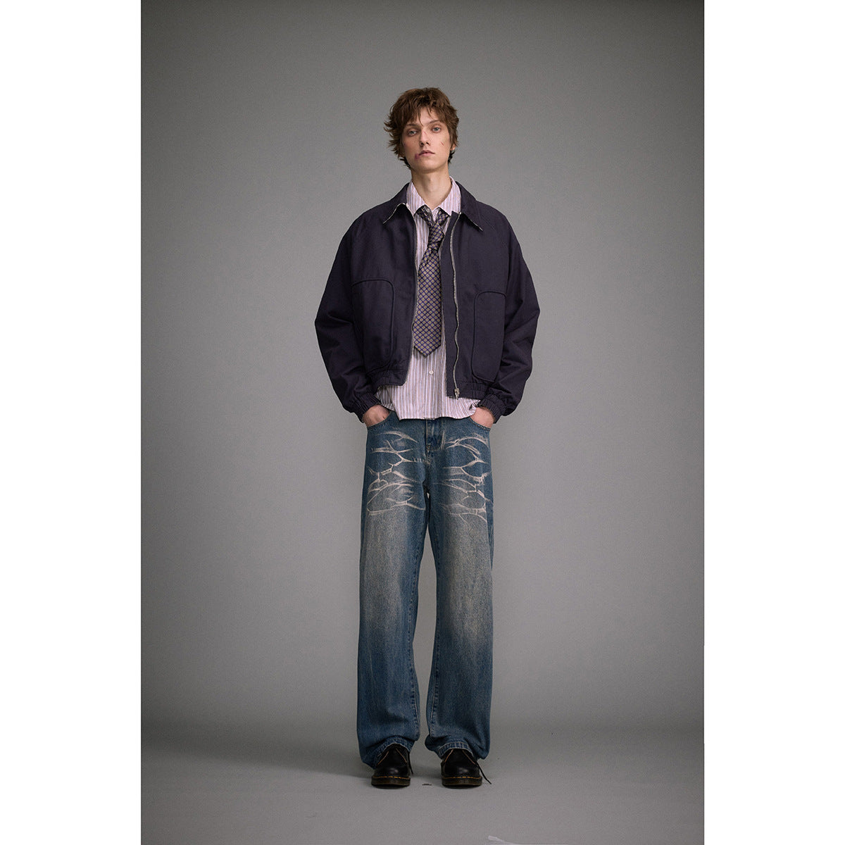 Front view of a model wearing loose blue jeans with pale abstract thigh designs, a navy jacket and tie.