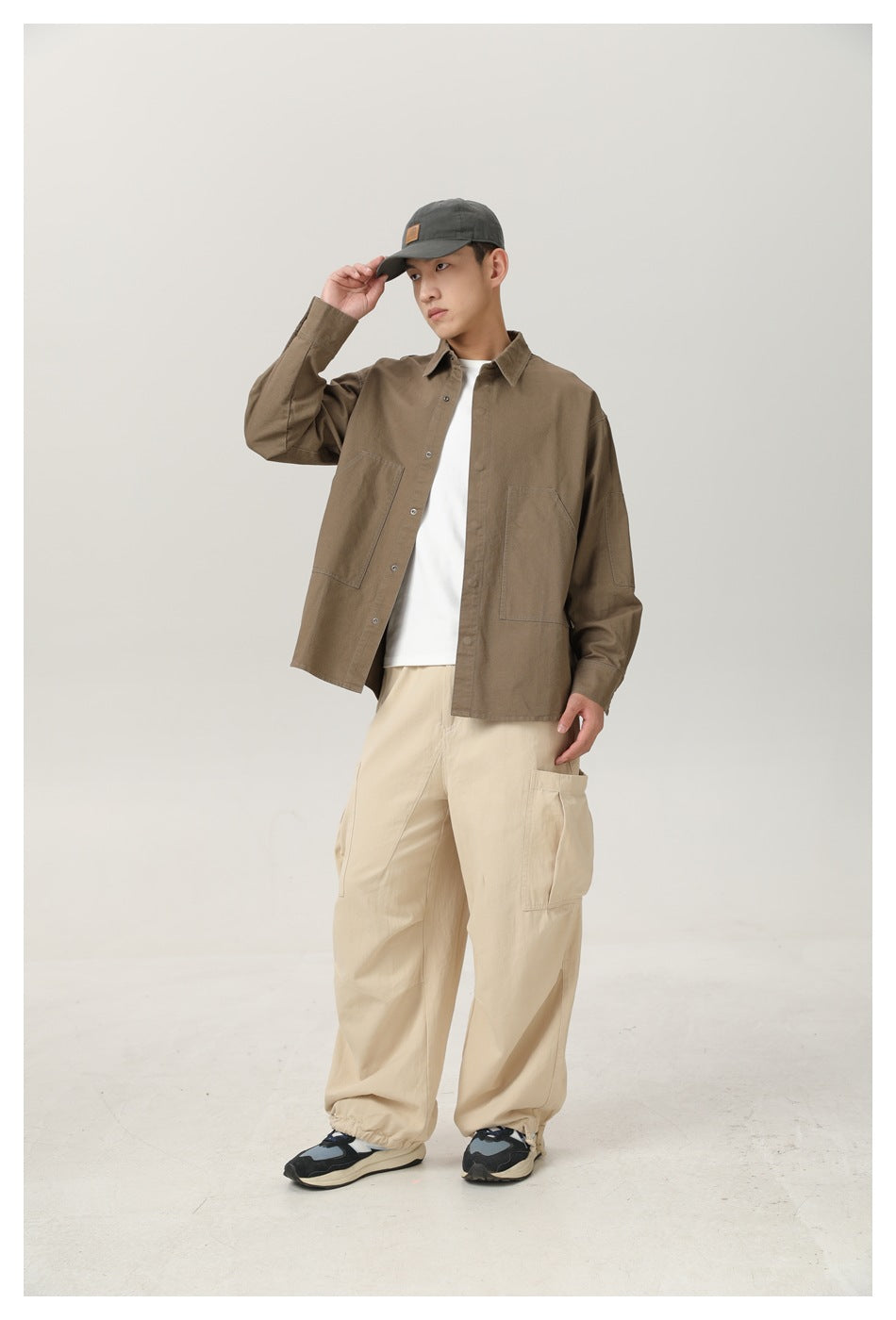 Full-length front view of a model wearing a brown pocketed overshirt, cream cargo pants and a dark cap.
