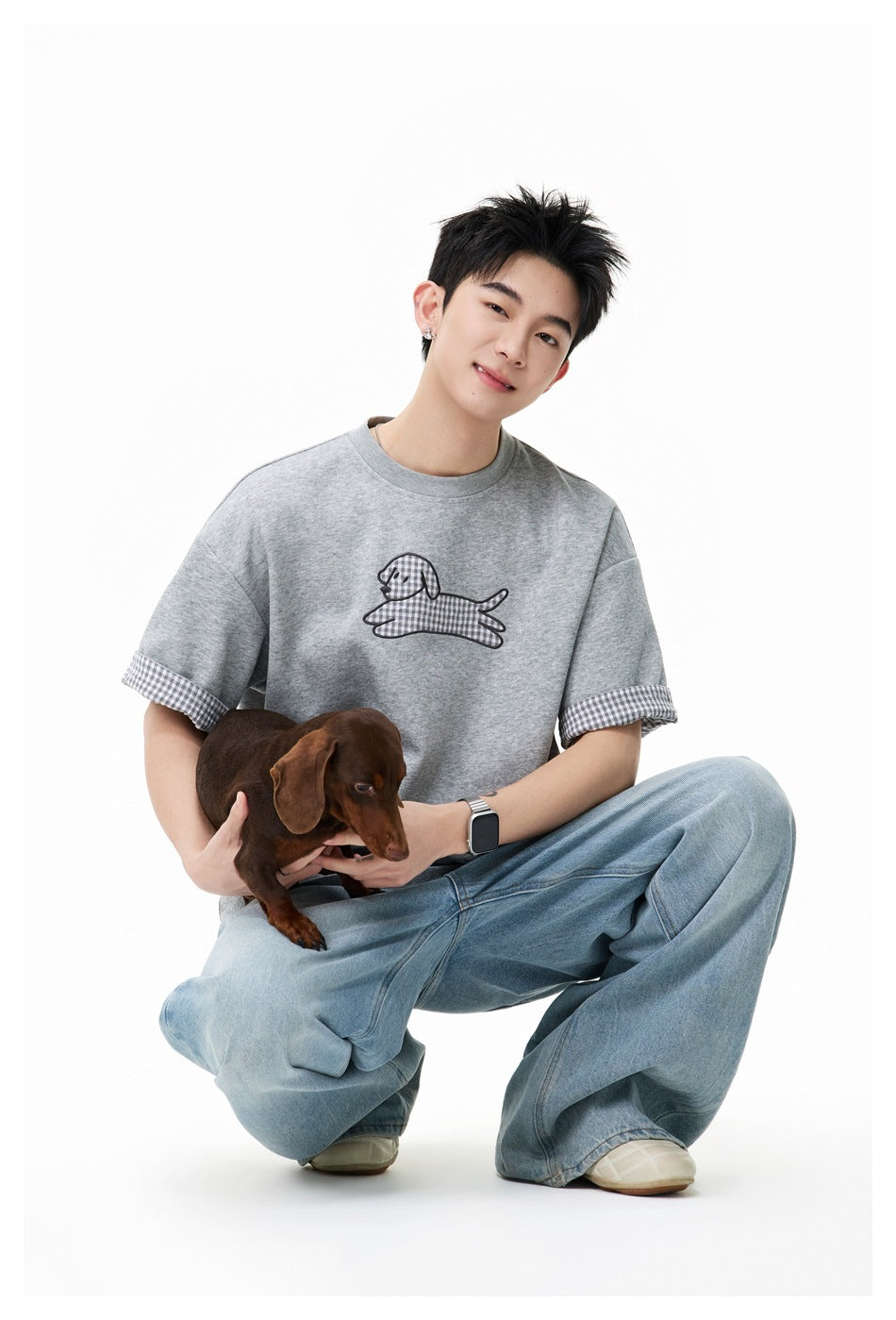Model crouching with a dachshund, wearing a gray T-shirt with a checked dog graphic and checked sleeve cuffs.