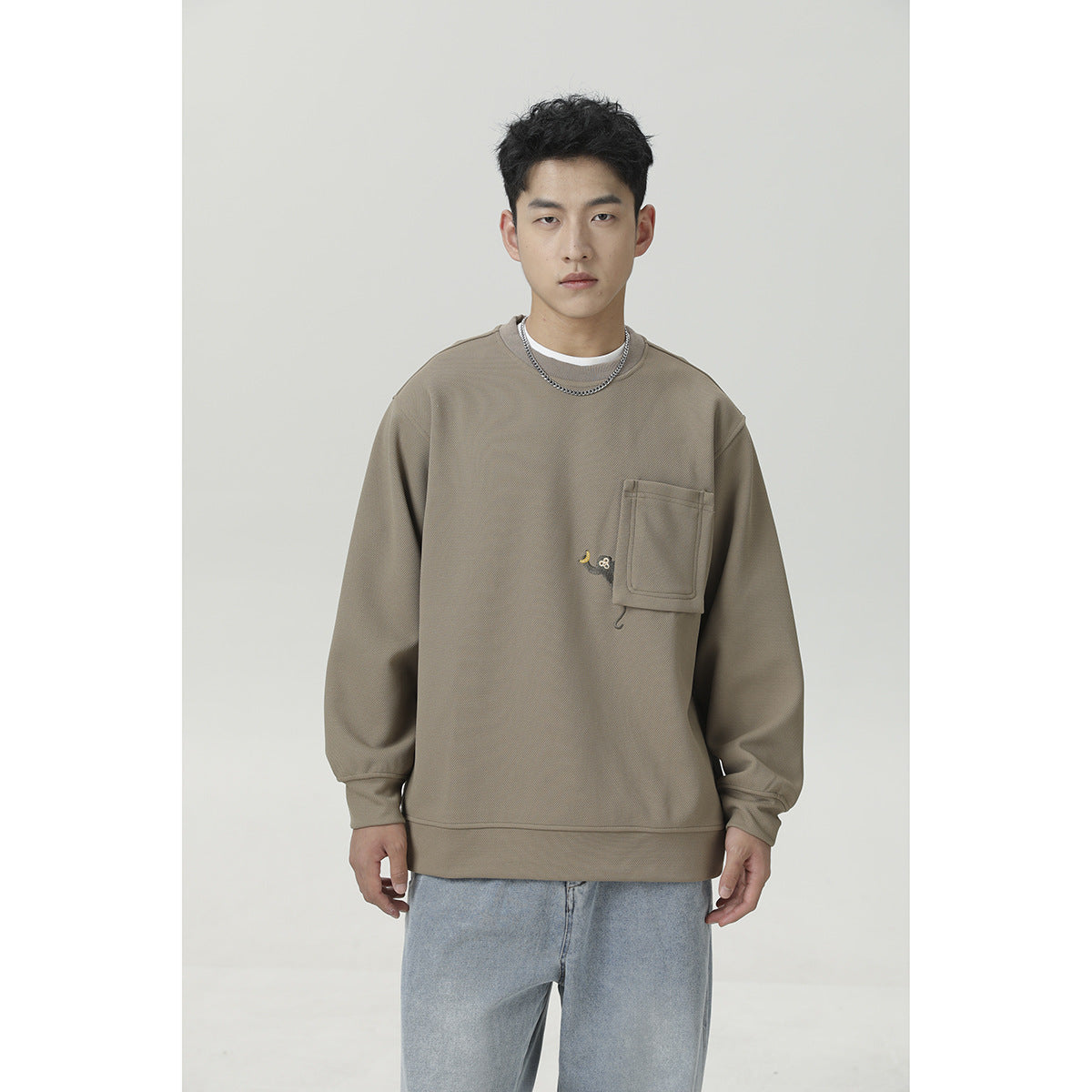 Front view of a man wearing a taupe crewneck sweatshirt with a chest pocket and light blue jeans.