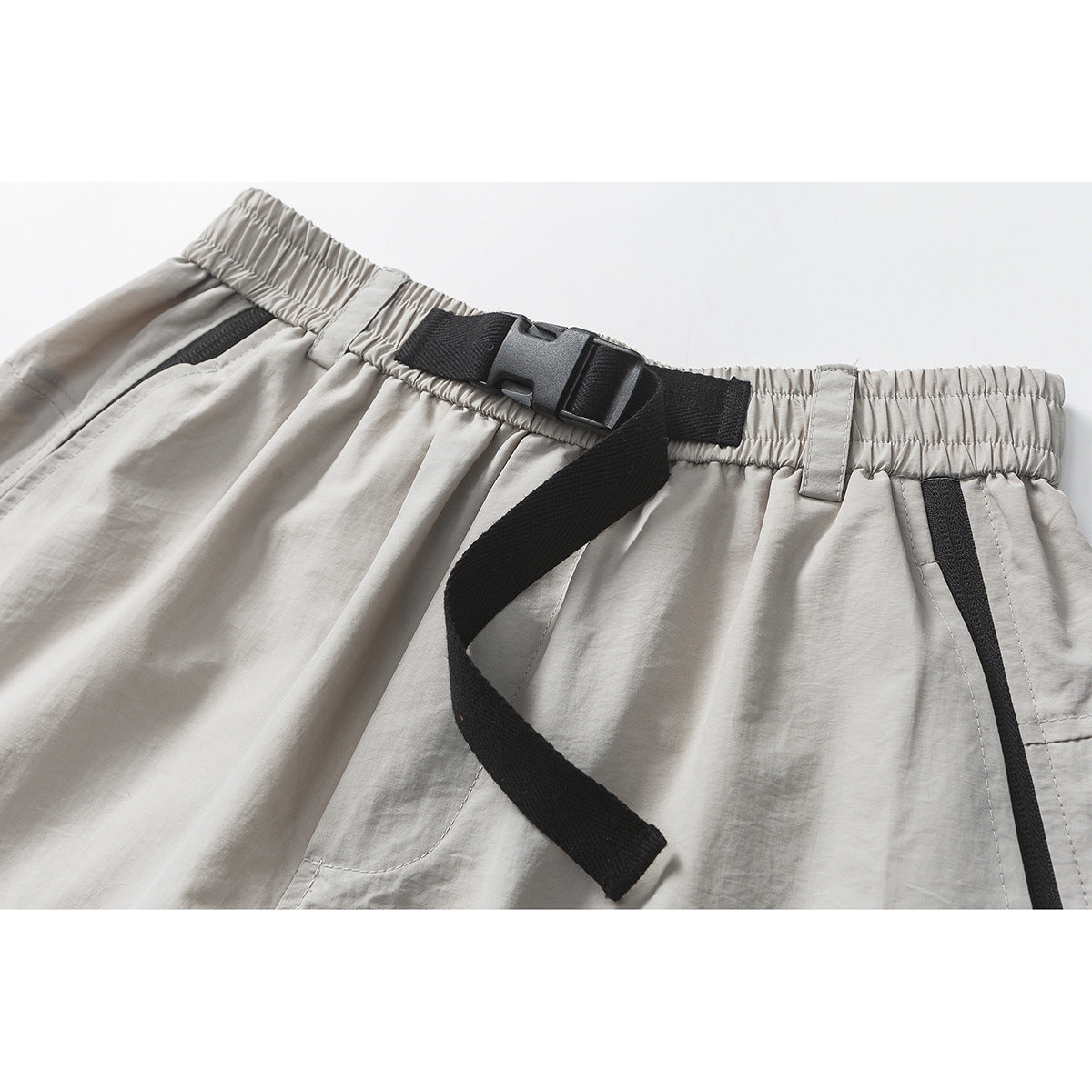 Close-up of light gray trousers with a gathered waistband, black buckle belt, and black-trimmed pocket.