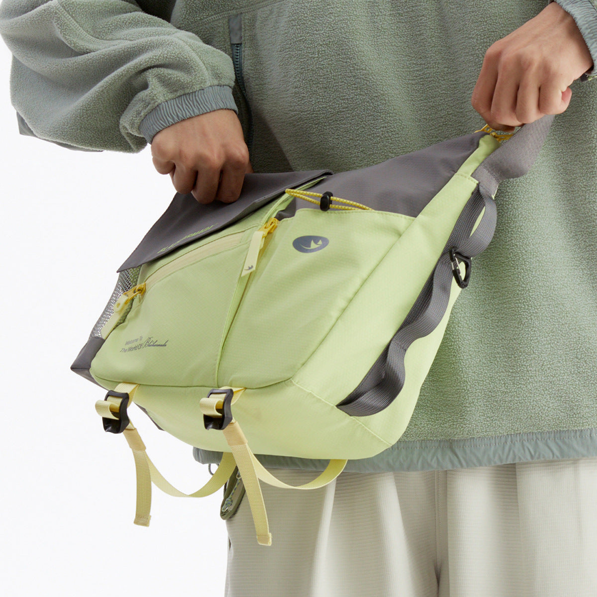 A person opens a pale lime and gray messenger bag, revealing its zip compartments and adjustable straps.