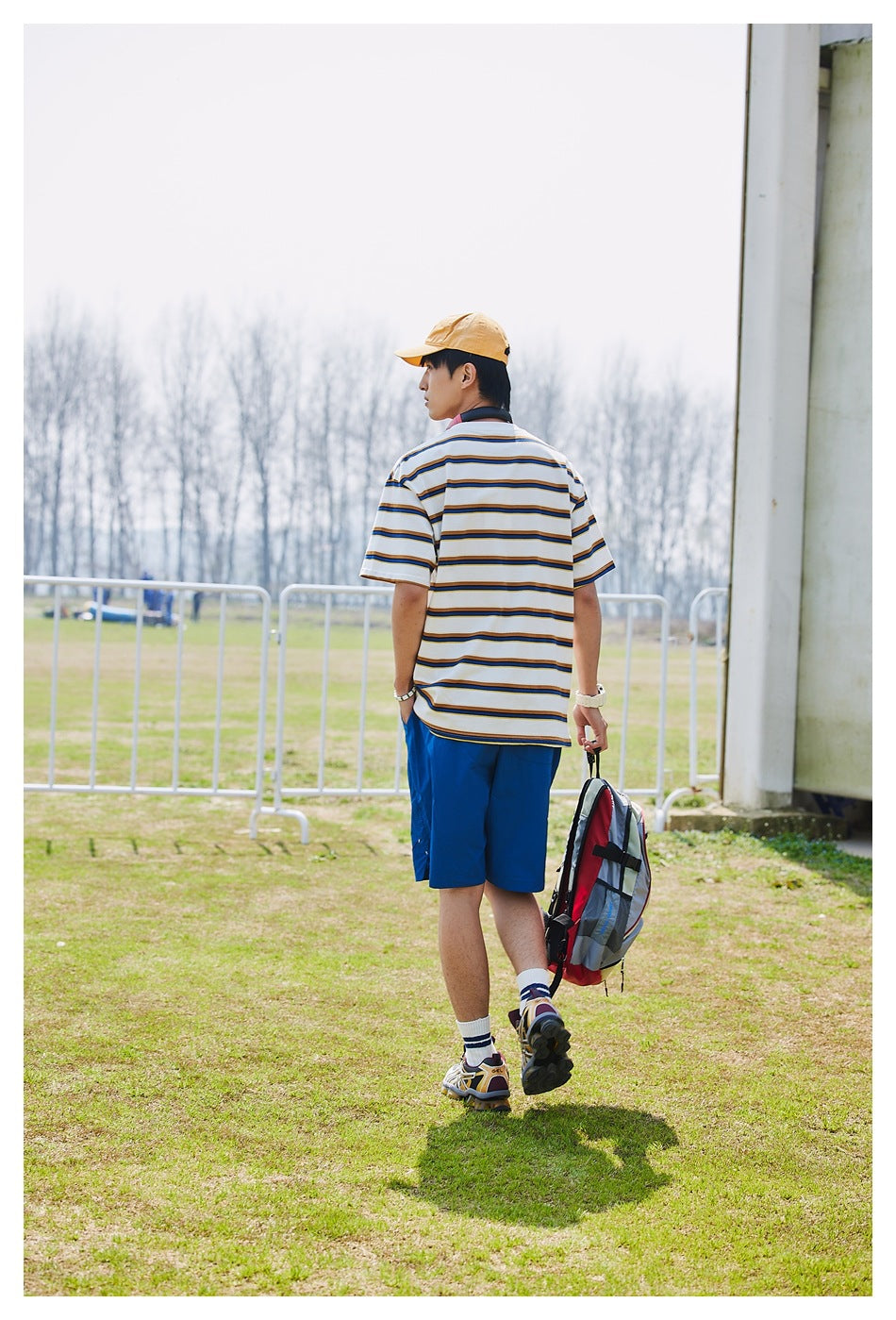 Rear view of a model wearing a white striped T-shirt and bright blue shorts while carrying a backpack.