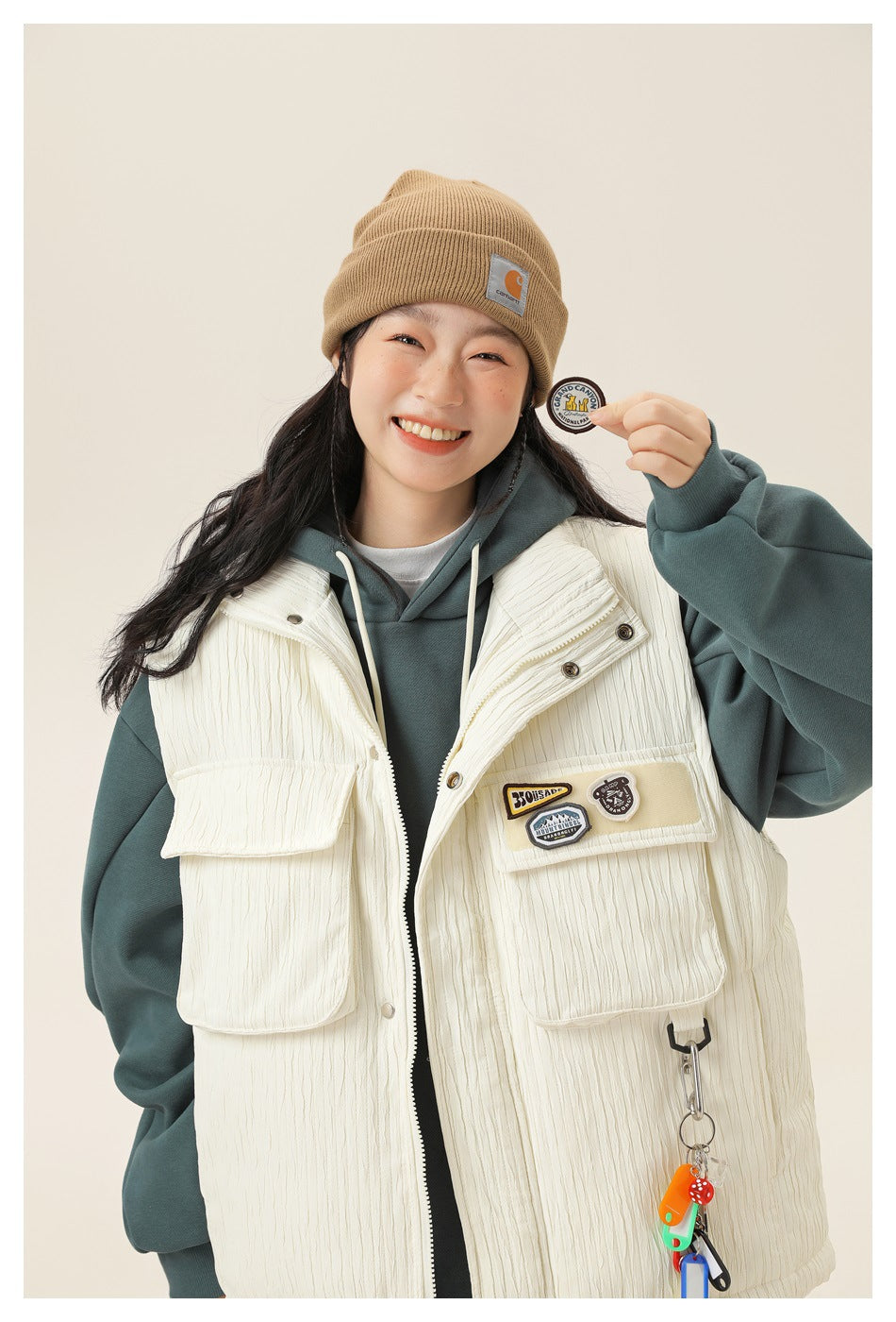 Close view of a model wearing a cream utility vest over a teal hoodie, showing the vest's flap pockets and patches.