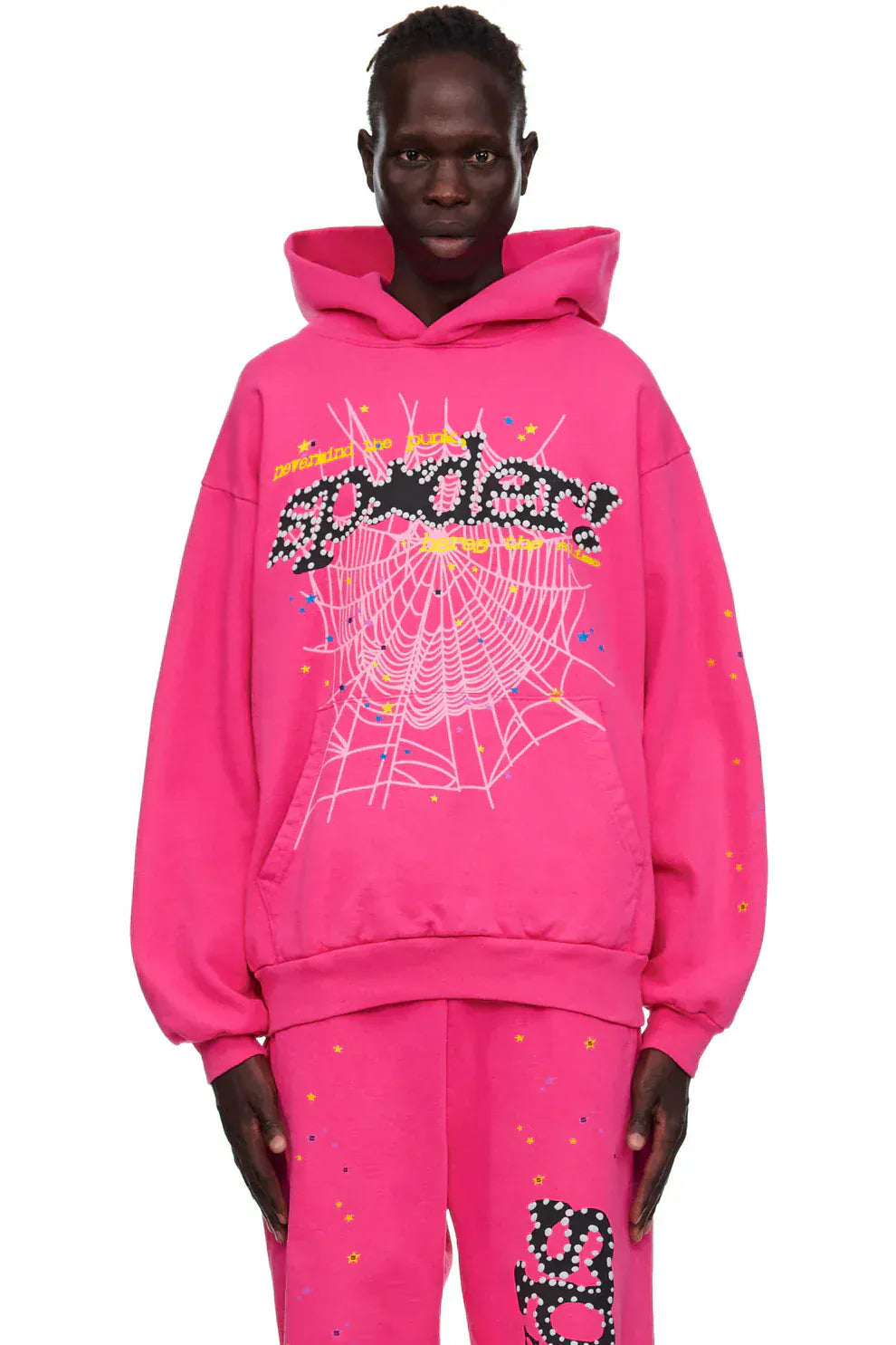 Wearing a Sp5der “Nevermind the Punk” Hoodie in Hot Pink (440GSM heavyweight streetwear pullover) with matching pants, featuring bold spiderweb and text graphics, a person stands confidently against a white background.