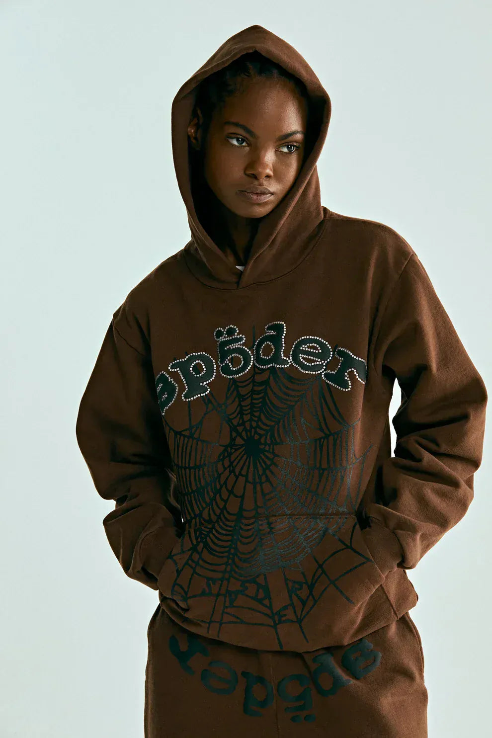 A person wearing a Sp5der Web Hoodie – Mocha Brown 440GSM Heavyweight Streetwear Pullover and matching pants, featuring a spider web and “spæder” printed on the front.