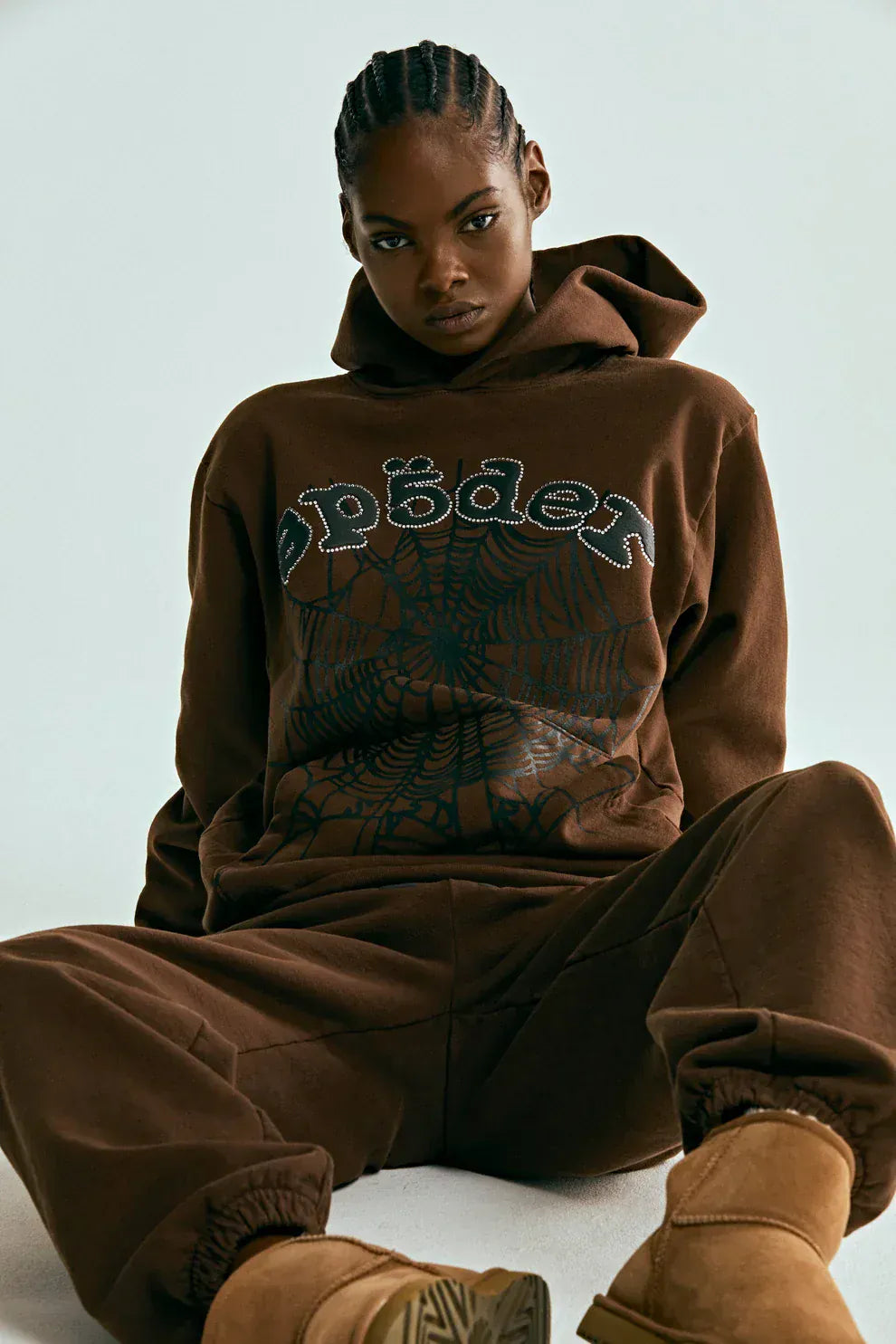 Person wearing a Sp5der Web Hoodie – Mocha Brown 440GSM Heavyweight Streetwear Pullover, paired with matching brown pants and tan boots, seated on the floor against a plain background.