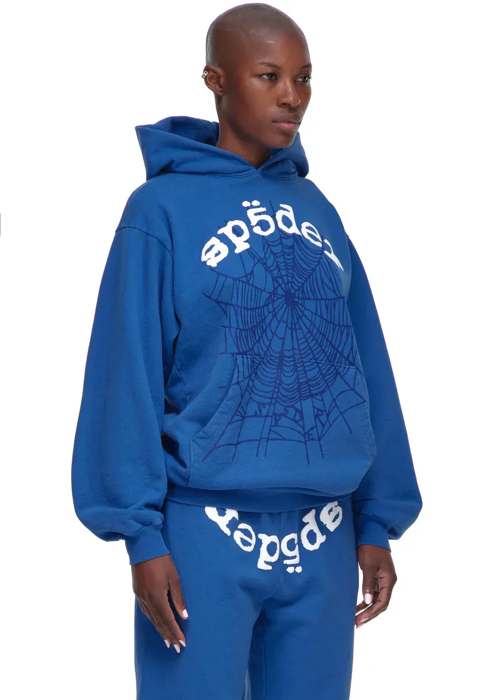 A person wearing the Sp5der OG Web Hoodie 440GSM in royal blue, a heavyweight cotton fleece sweatshirt with a white spider-web graphic and “sp5der” print, styled with matching royal-blue sweatpants.