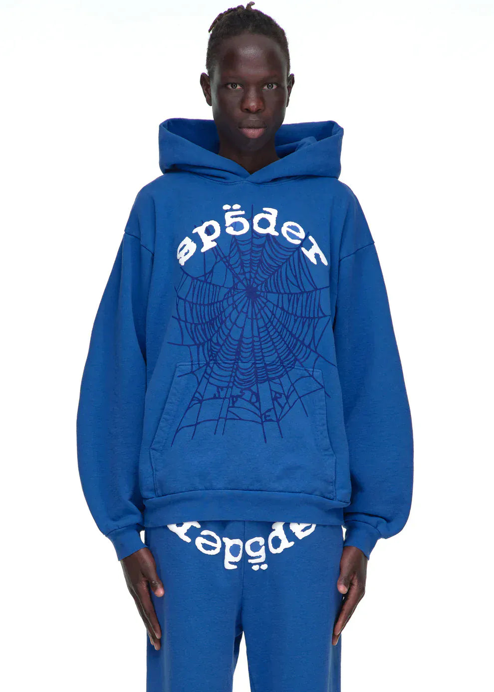 Someone models the Sp5der OG Web Hoodie 440GSM, a royal blue heavyweight sweatshirt featuring white text and a spider-web graphic, standing against a plain white background.