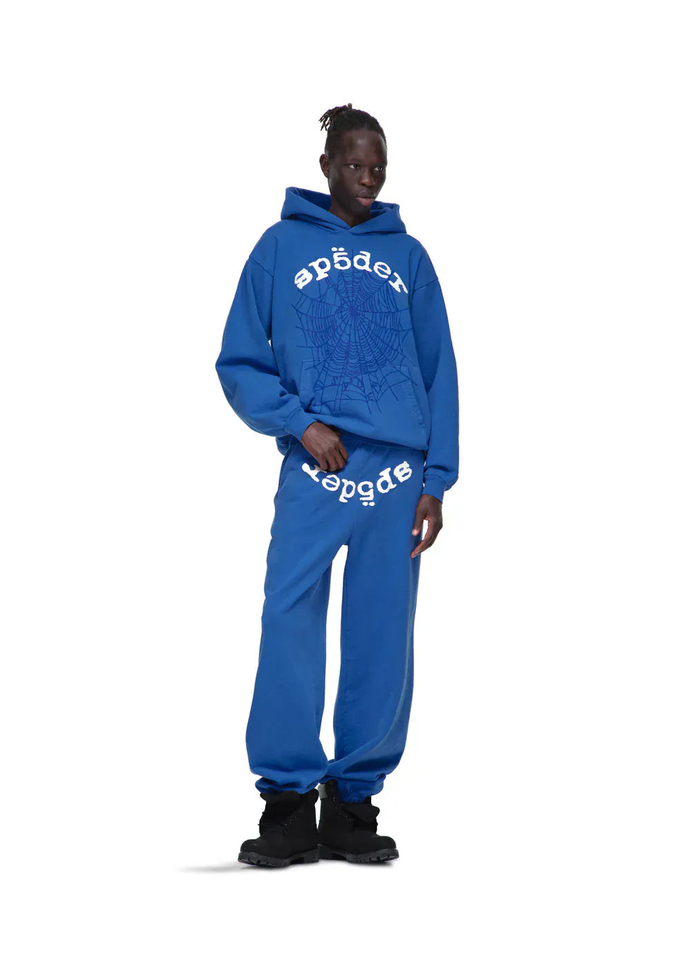 A person in a Sp5der OG Web Hoodie 440GSM – Royal Blue Heavyweight Spider-Web Graphic Sweatshirt and matching blue sweatpants with white web graphics and text, paired with black shoes, stands against a plain white background.