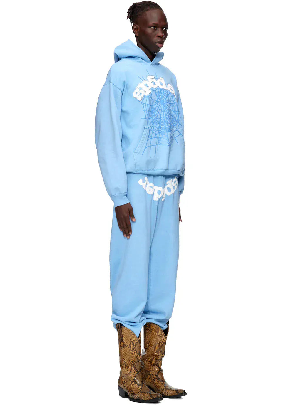 A person stands in profile wearing a Sp5der Hoodie – Sky Blue 440GSM Heavyweight Chenille Logo Spider-Web Streetwear Pullover and matching sweatpants, accented with bold white graphics and tan snakeskin cowboy boots for a standout streetwear style.