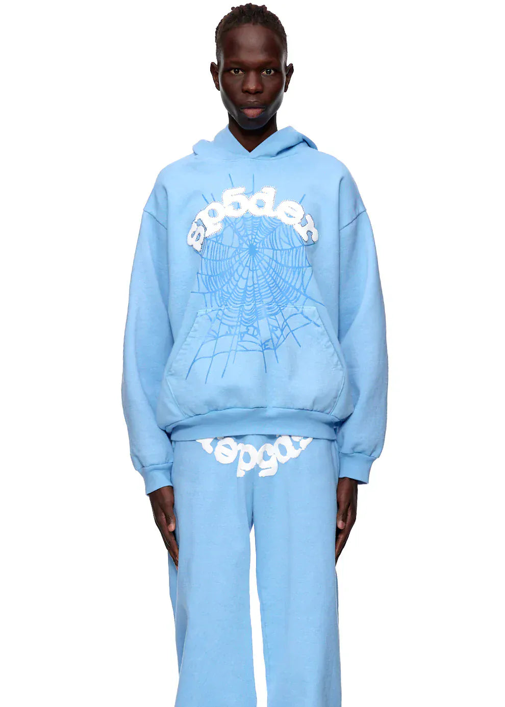 Wearing the Sp5der Hoodie – Sky Blue 440GSM Heavyweight Chenille Logo Spider-Web Streetwear Pullover and matching sweatpants with white graphics, a person stands against a plain white background for a clean streetwear look.