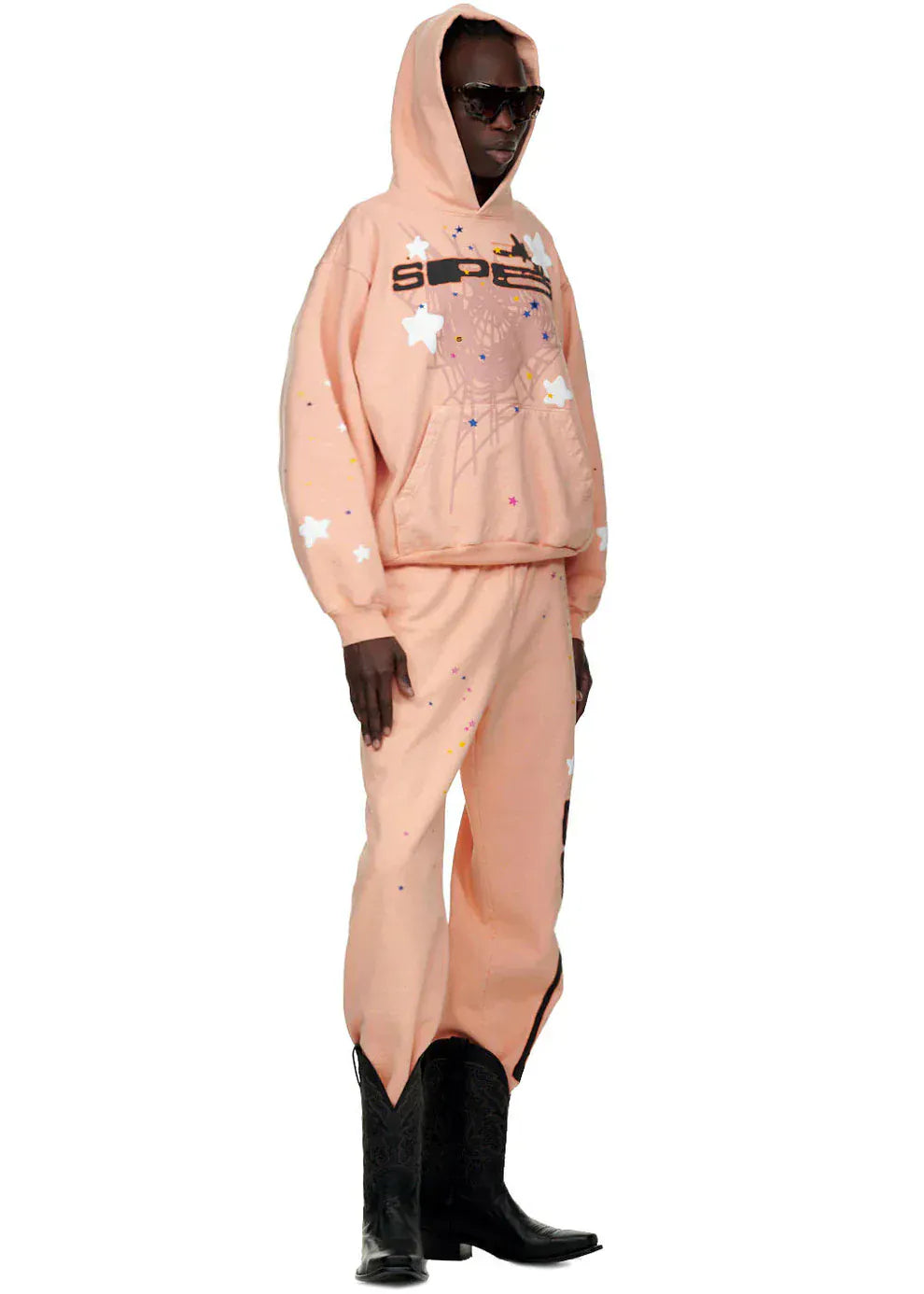 A person wears the Sp5der Star Web Hoodie – Peach Pink 440GSM Heavyweight Oversized Streetwear Pullover with matching star pants, black cowboy boots, and sunglasses, posing against a white background in true Y2K streetwear style.