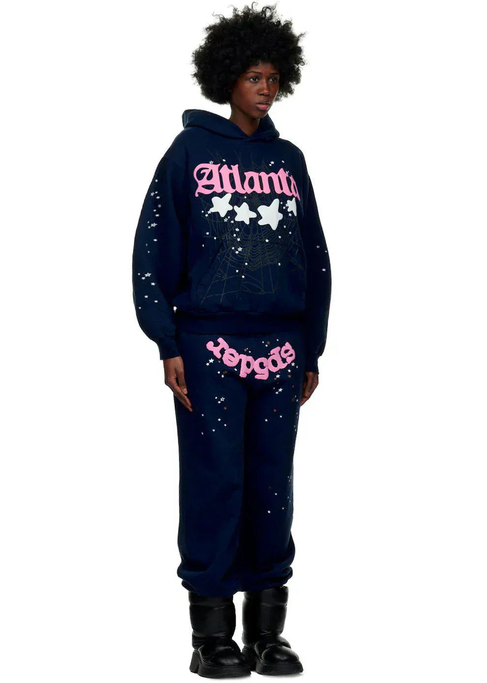 A person wears the Sp5der “Atlanta Nights” Web Hoodie – Navy Blue 440GSM Heavyweight Oversized Streetwear Pullover, paired with sweatpants featuring pink and white graphic text, star designs, and black boots against a plain white background.