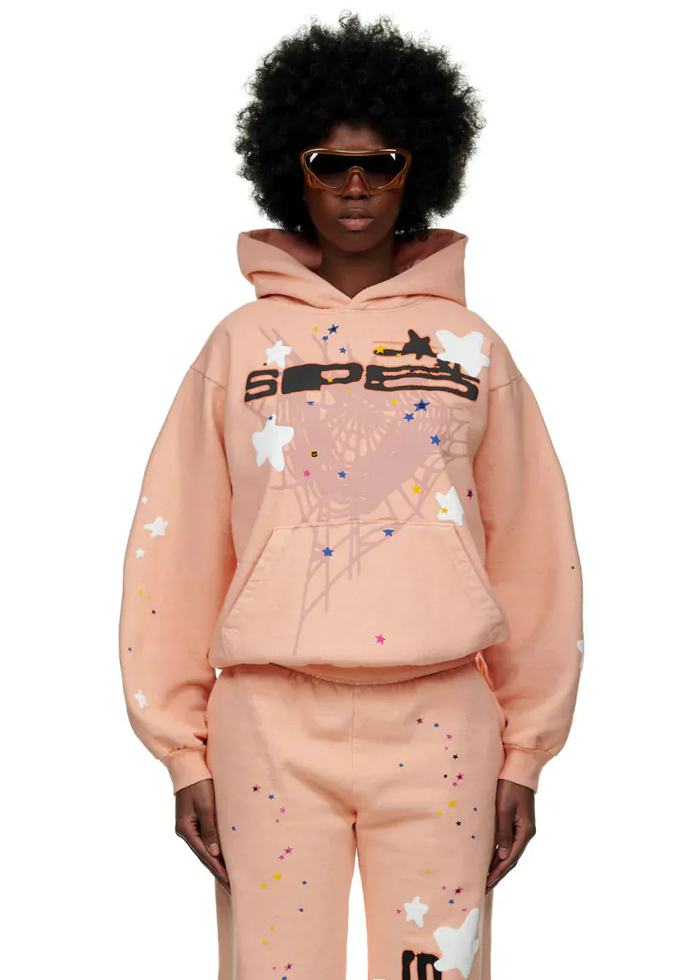 A person wearing sunglasses and a Sp5der Star Web Hoodie – Peach Pink 440GSM Heavyweight Oversized Streetwear Pullover with matching sweatpants stands against a plain background.