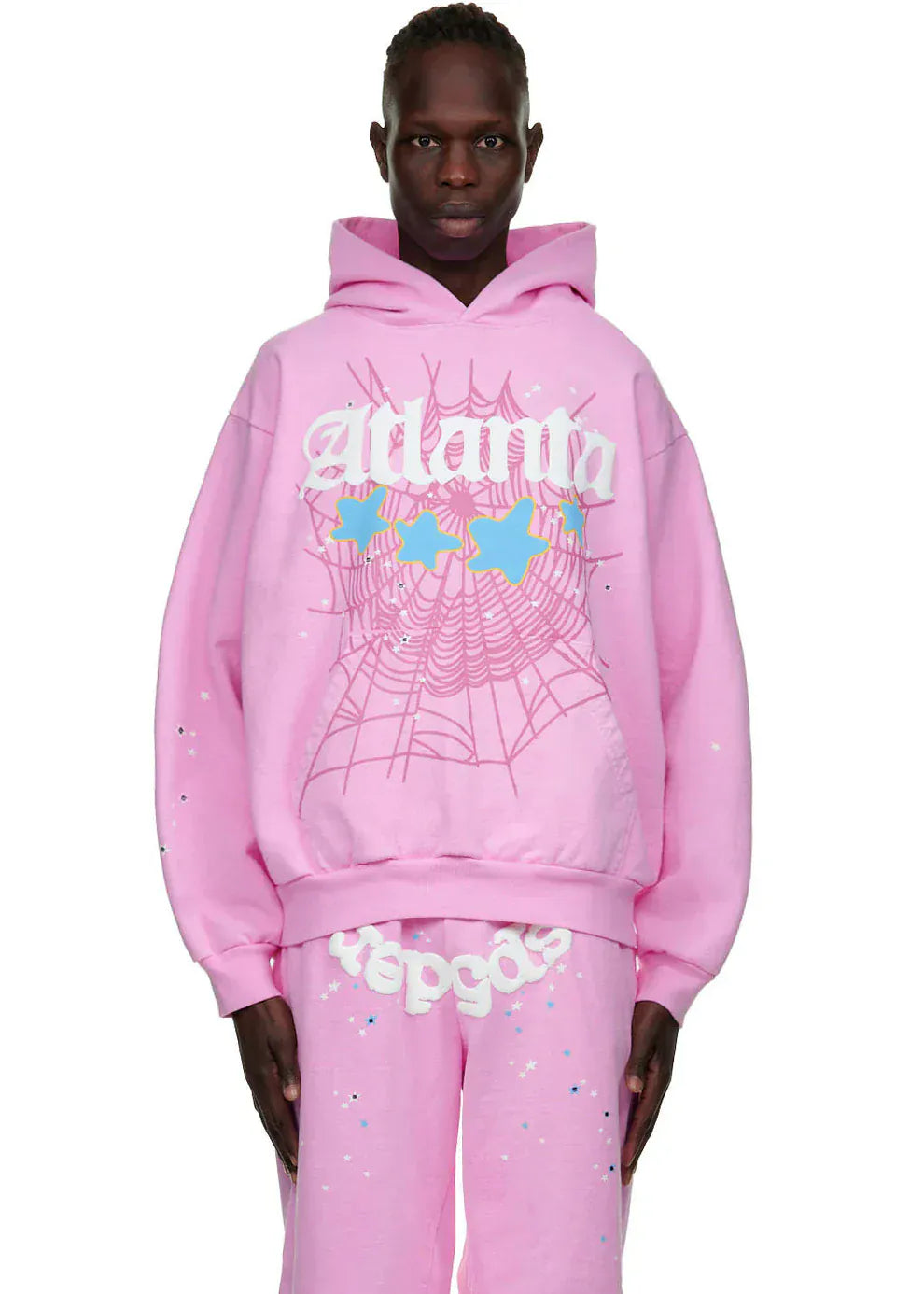 A person wears the Sp5der Atlanta Hoodie – Pink 440GSM Heavyweight Y2K Streetwear Pullover with matching graphic pants, featuring a spider web and star design, standing against a white background.