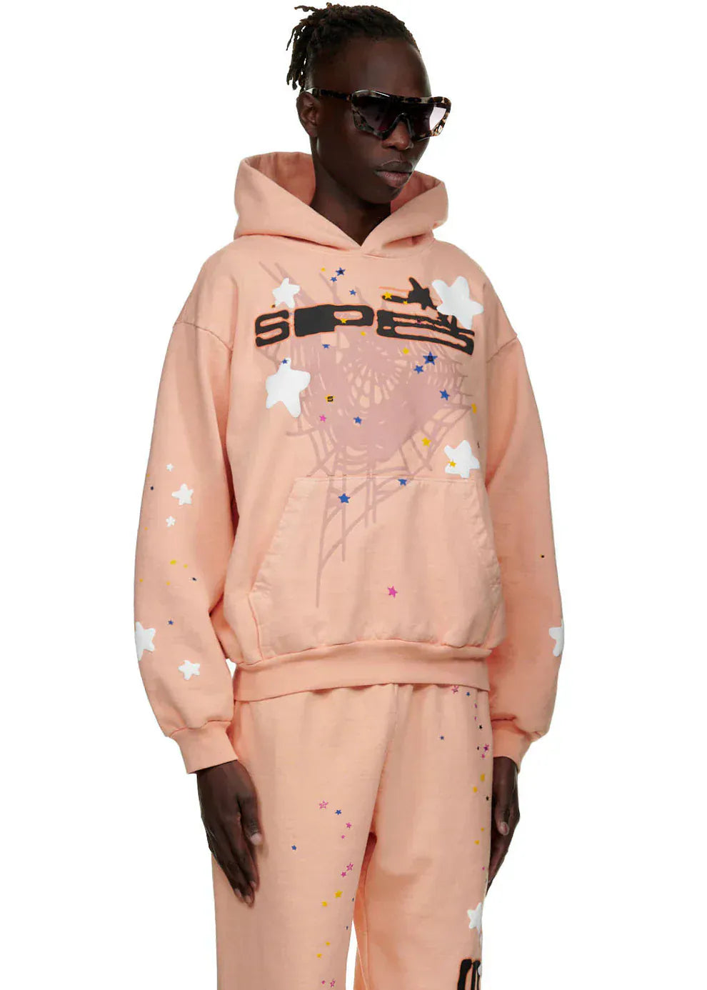 A person wearing the Sp5der Star Web Hoodie – Peach Pink 440GSM Heavyweight Oversized Streetwear Pullover and matching sweatpants with star graphics stands against a plain background, embodying Y2K streetwear vibes.