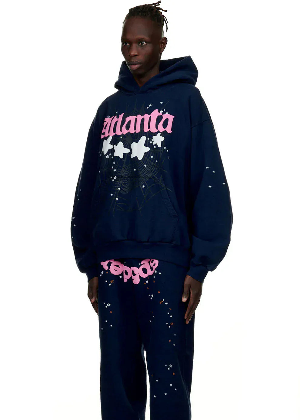 Dressed in a Sp5der “Atlanta Nights” Web Hoodie—navy blue, 440GSM heavyweight oversized streetwear pullover with white splatters, pink text, and stars—a person stands against a plain white background.