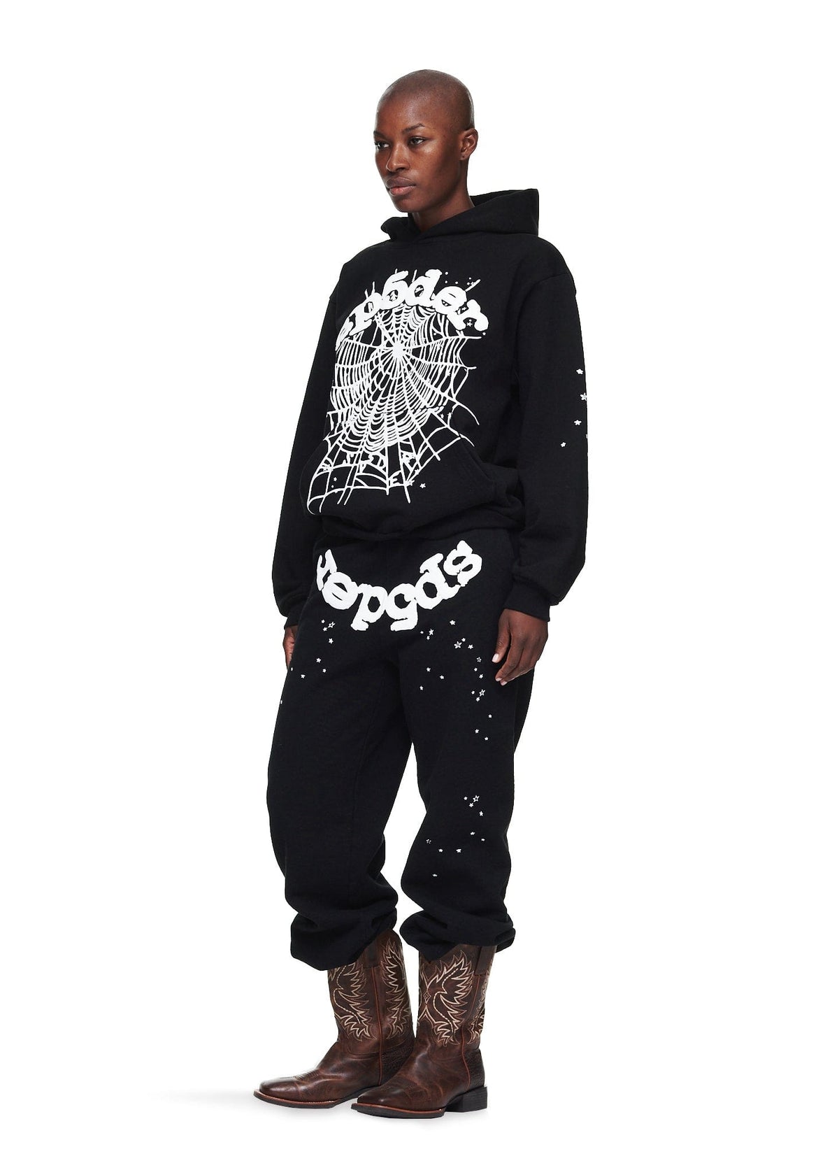 Individual dressed in a Sp5der “Webcore Noir” Hoodie – Black & White 440GSM Heavyweight Oversized Streetwear Pullover and matching sweatpants with white graphics, paired with brown cowboy boots, stands against a white background.