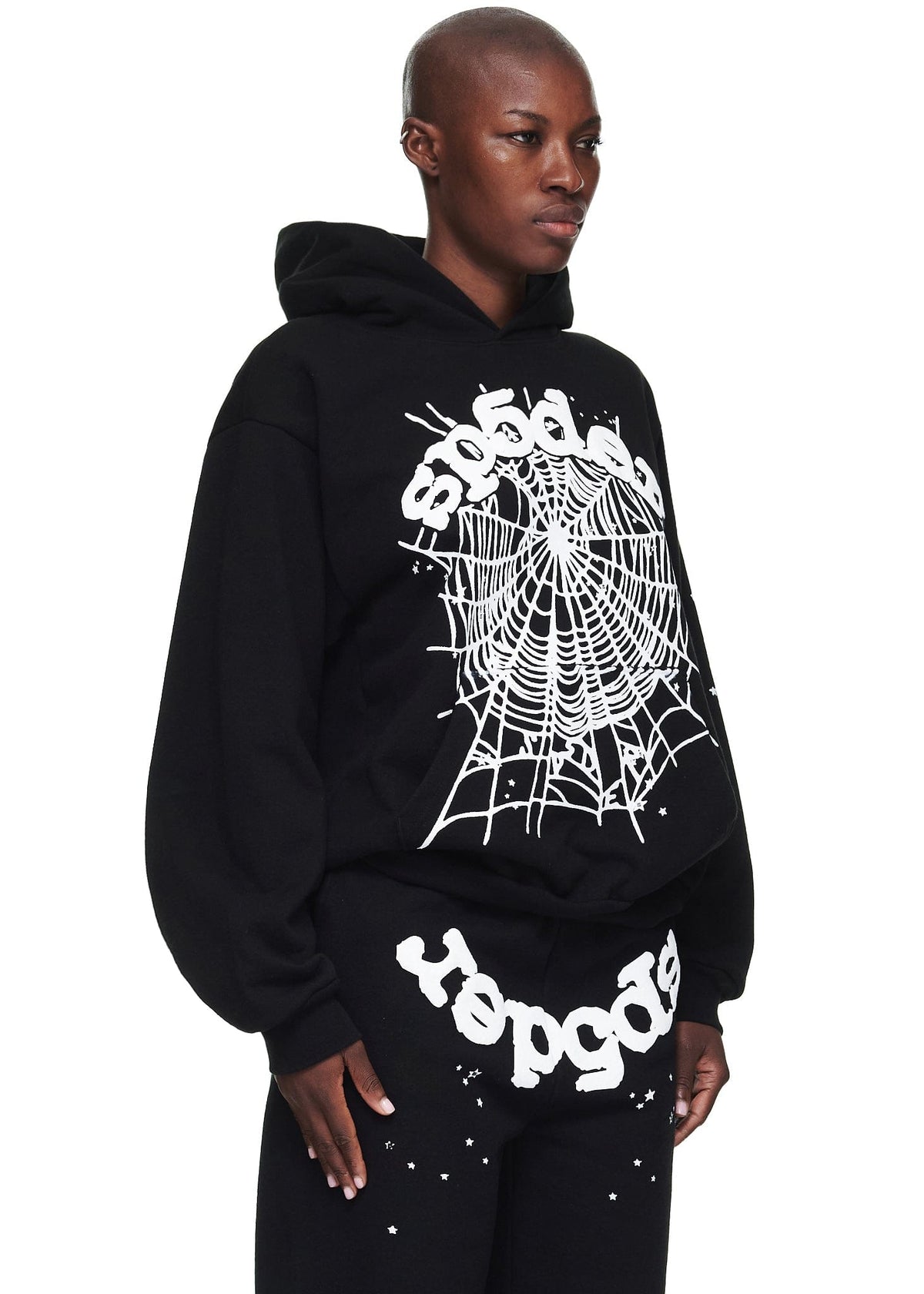 Someone wears the Sp5der “Webcore Noir” Hoodie – a black & white 440GSM heavyweight oversized streetwear pullover with spiderweb designs, paired with matching pants, standing against a plain white background.