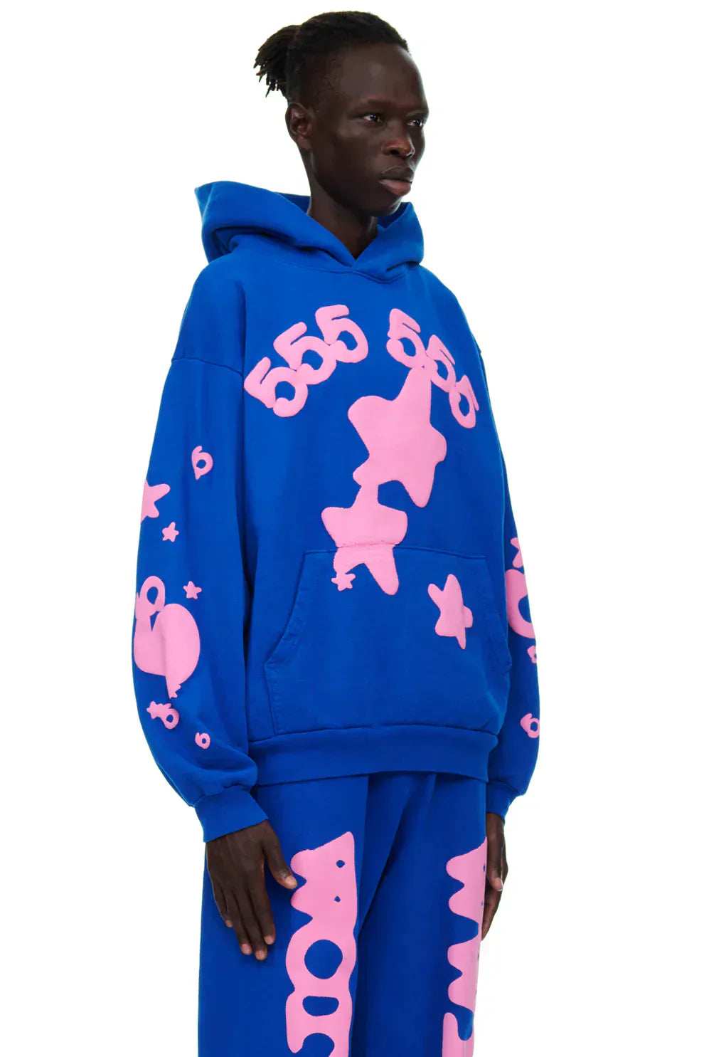 A person in a Sp5der “555 Stars” Hoodie – Royal Blue/Pink 440GSM Heavyweight Oversized Streetwear Pullover with matching pants featuring bold pink graphics, embodying Y2K streetwear, stands against a white background.