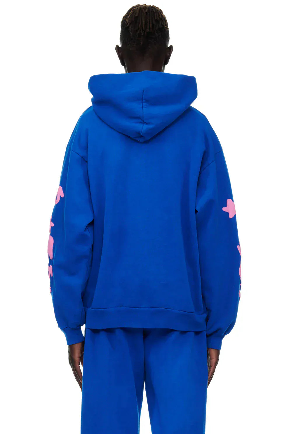 Facing away, a person wears the Sp5der “555 Stars” Hoodie in royal blue/pink—an oversized streetwear pullover made from 440GSM heavyweight cotton fleece—with matching blue pants, capturing a bold Y2K streetwear vibe.