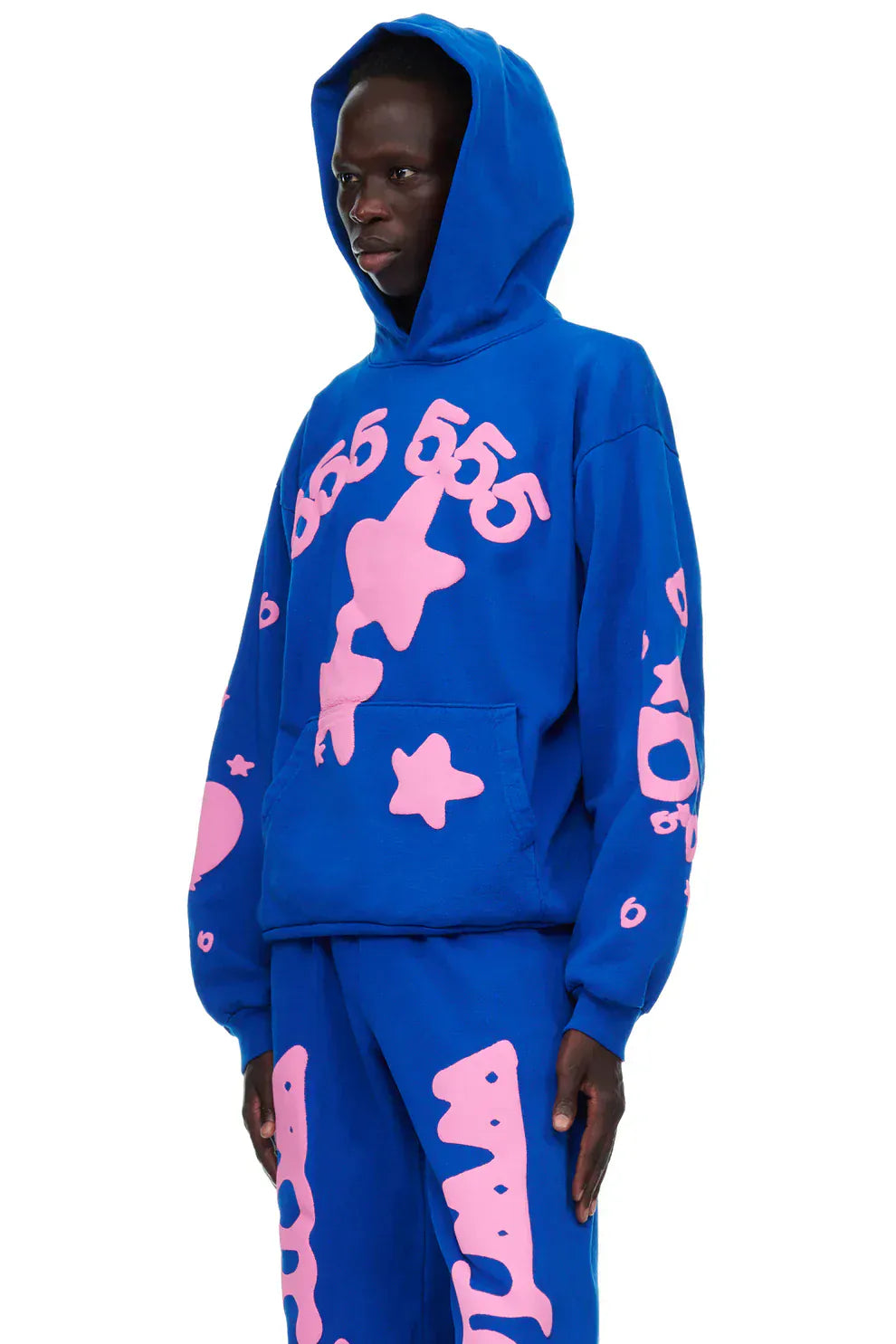 Person wearing the Sp5der “555 Stars” Hoodie in royal blue/pink (440GSM heavyweight oversized streetwear pullover) with matching pants featuring bold pink graphics and numbers, standing against a white background.
