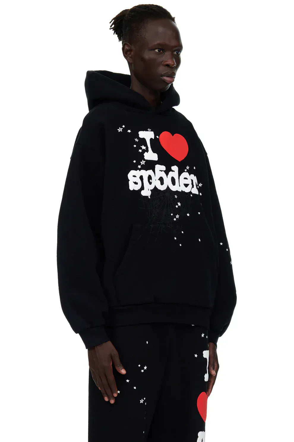 Wearing the Sp5der “I Love Sp5der” Hoodie – Black Web 440GSM Heavyweight Pullover and matching star pants, a person showcases bold streetwear style against a plain background.