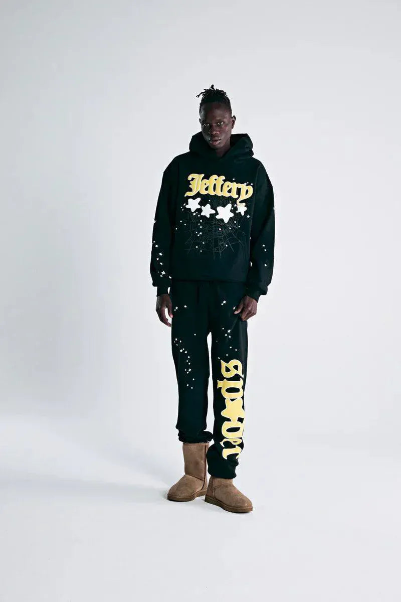 A person stands against a plain background wearing the Sp5der Jeffery Hoodie – Black Web, a 440GSM heavyweight streetwear pullover, paired with sweatpants featuring yellow lettering and tan boots.