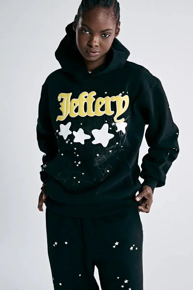 A person wears the Sp5der Jeffery Hoodie – Black Web 440GSM Heavyweight Streetwear Pullover with yellow text, white stars, and a spiderweb design, paired with black pants featuring scattered white stars for a standout streetwear look.