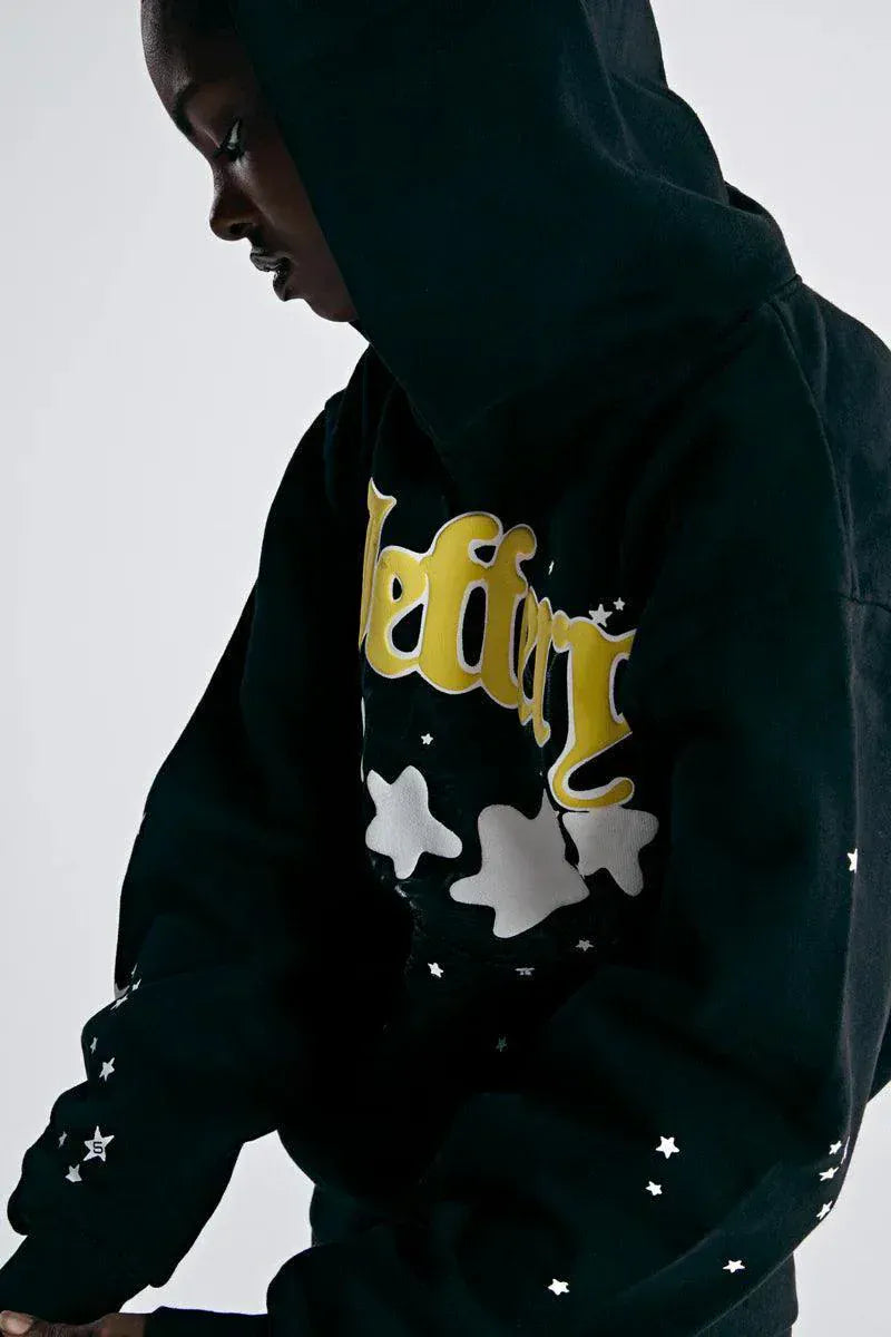 A person in a Sp5der Jeffery Hoodie – Black Web 440GSM Heavyweight Streetwear Pullover with yellow lettering and white star and cloud designs stands sideways against a plain background.