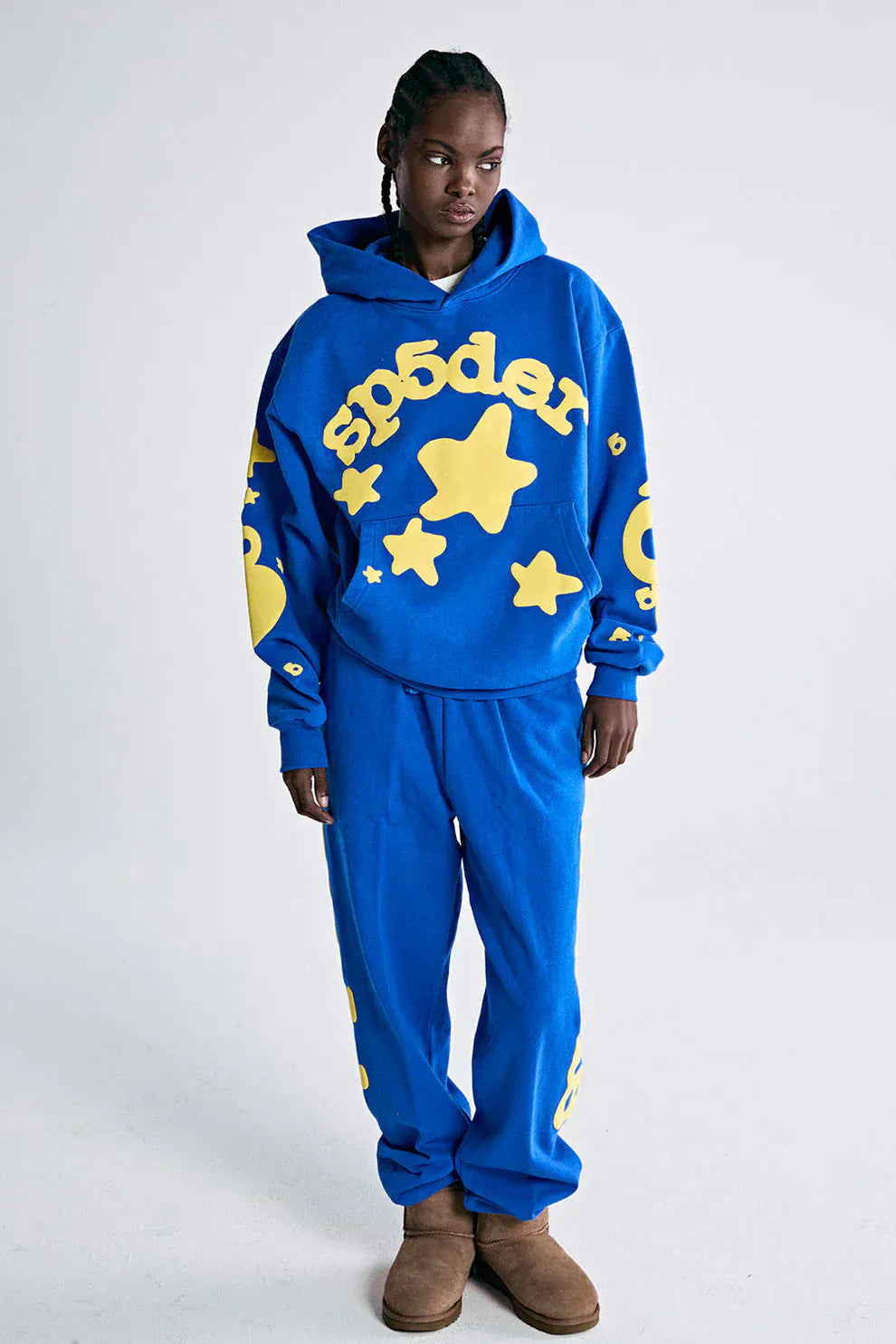 Wearing the Sp5der 440GSM Heavyweight Hoodie in royal blue with yellow star 3D puff print, this look channels Y2K streetwear against a clean white background, paired with matching sweatpants and comfy brown slippers.