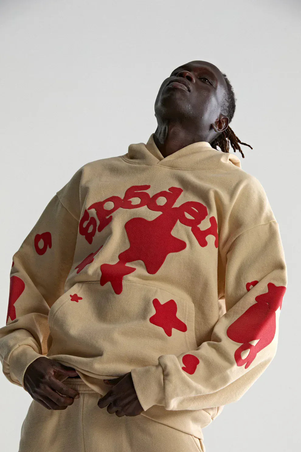 Someone poses against a light background in the Sp5der 440GSM Heavyweight Star Hoodie—a cream oversized streetwear pullover featuring red puff print star designs.