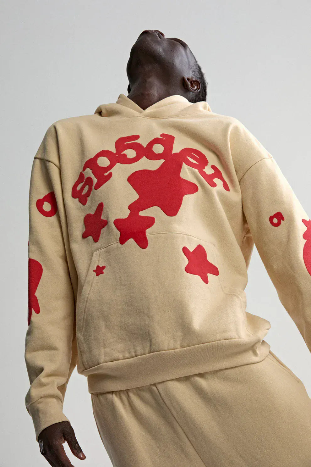 Model wearing a Sp5der 440GSM Heavyweight Star Hoodie in cream with red puff print, styled with matching pants, leaning back and gazing upward—showcasing an oversized streetwear pullover against a plain backdrop.