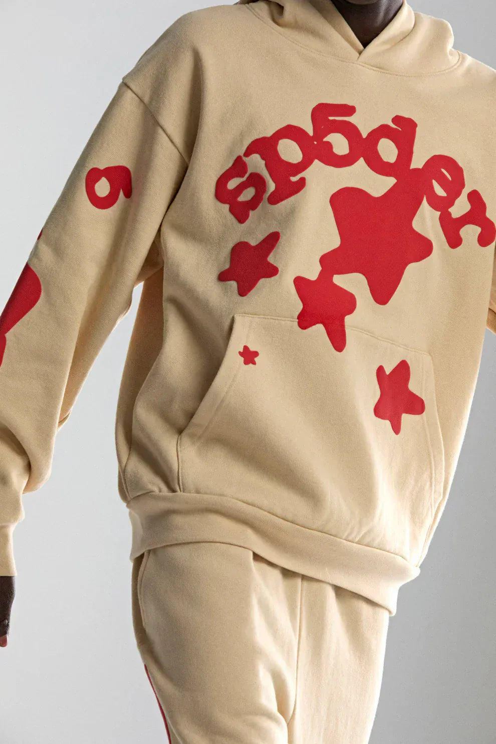 Someone wearing the Sp5der 440GSM Heavyweight Star Hoodie in cream with red puff print—a bold, oversized streetwear pullover featuring standout graphic stars and iconic Sp5der branding.