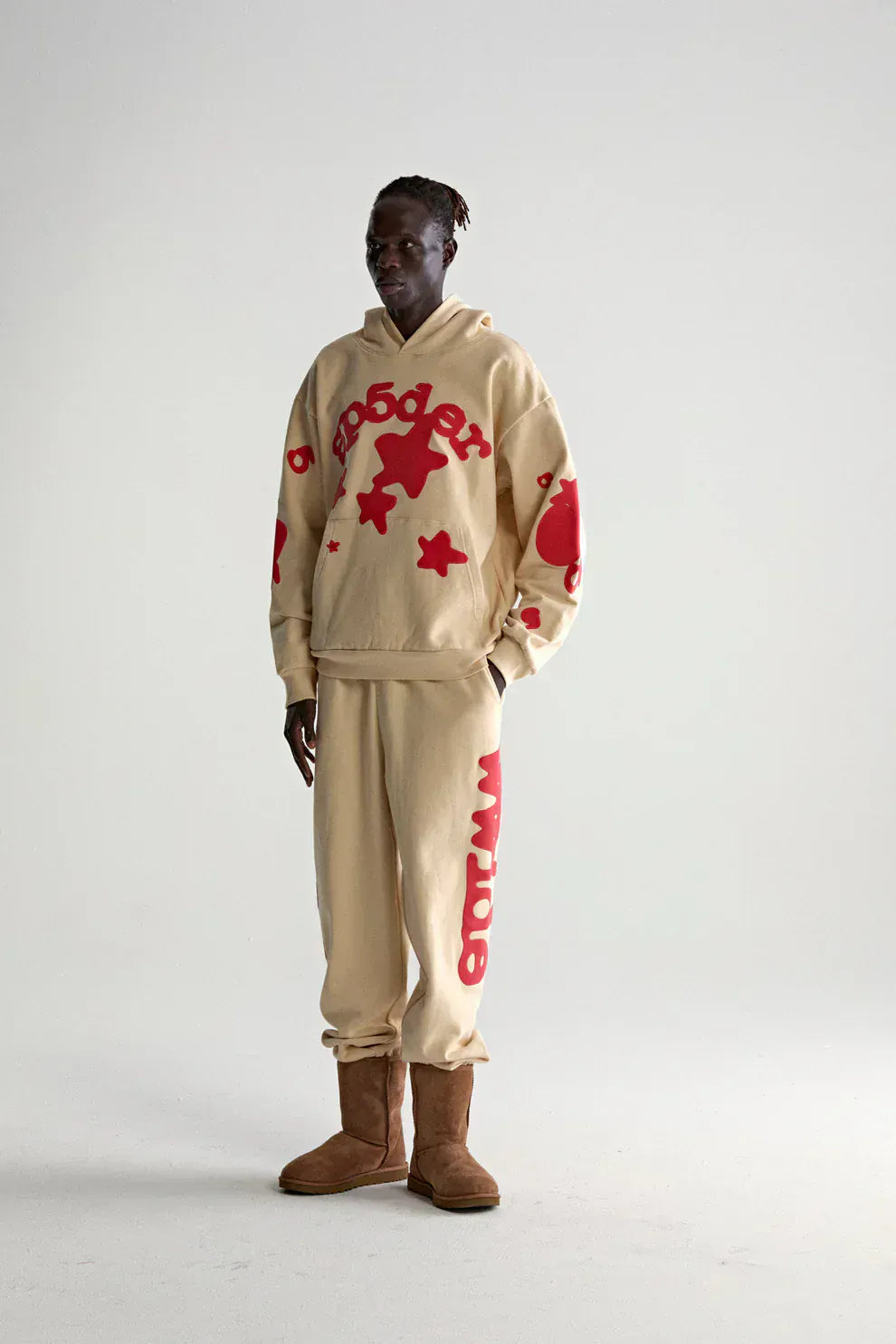 A person wears the Sp5der 440GSM Heavyweight Star Hoodie in cream with red puff print, styled as an oversized streetwear pullover, paired with sweatpants and brown boots, standing against a plain white background.